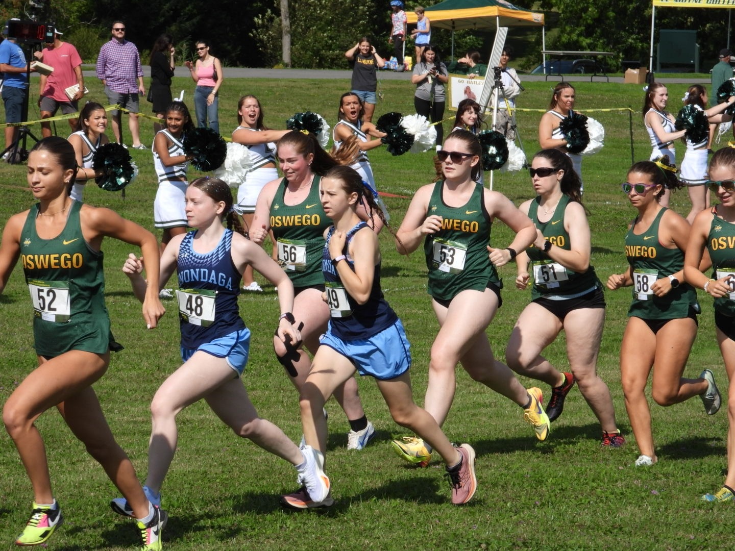 A women’s cross-country race is underway on a grassy course. Several runners in green Oswego uniforms and others in blue uniforms are competing in the foreground, with race officials, spectators, and cheerleaders visible behind them. The runners are wearing numbered race bibs, and a timing display and event tents can be seen in the background.