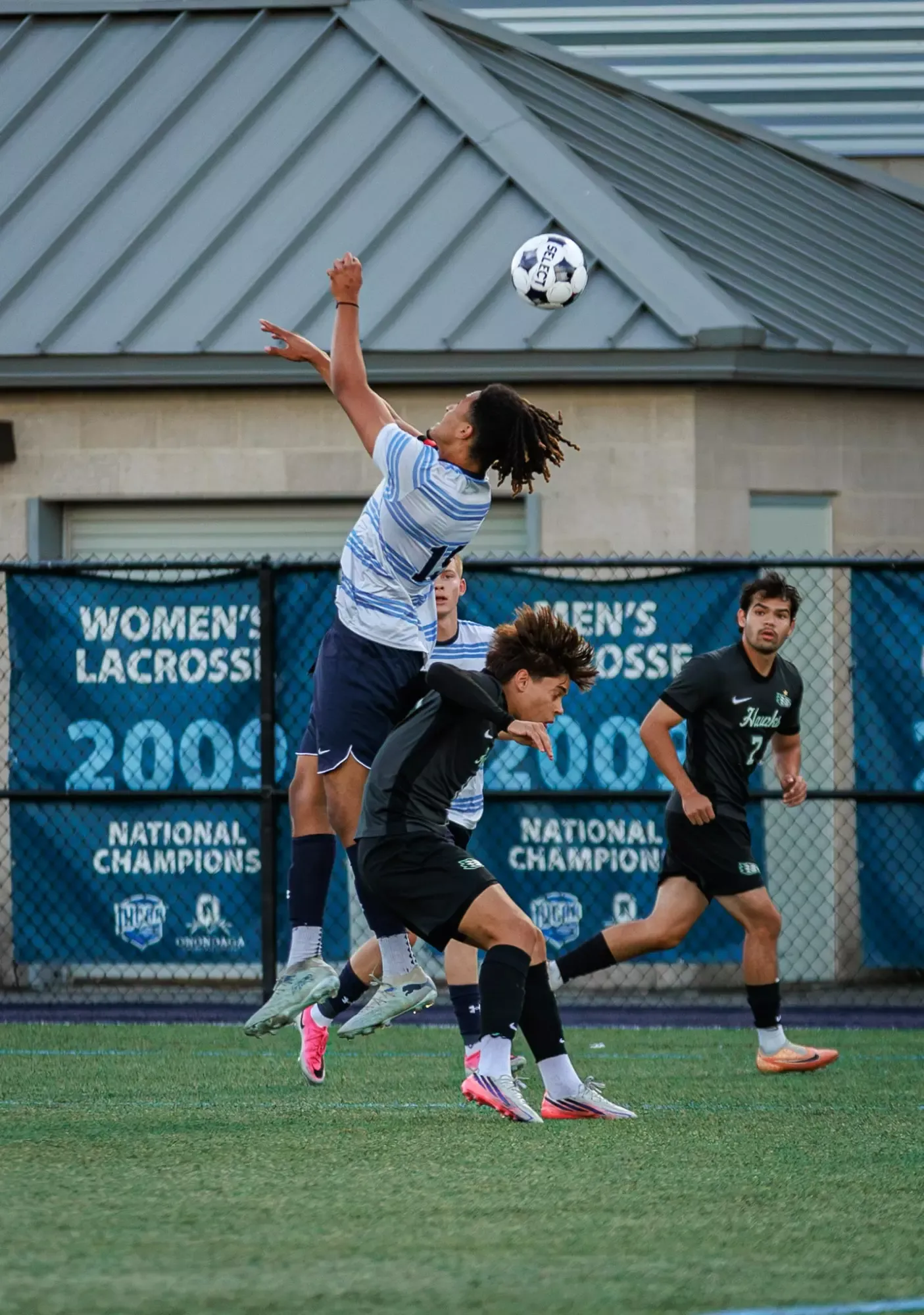 A soccer player leaps to head a ball during a match, with several other players running nearby on an outdoor artificial-turf field. A chain-link fence and a building with a metal roof are visible in the background. Signs behind the players reference women’s and men’s lacrosse and national championships.