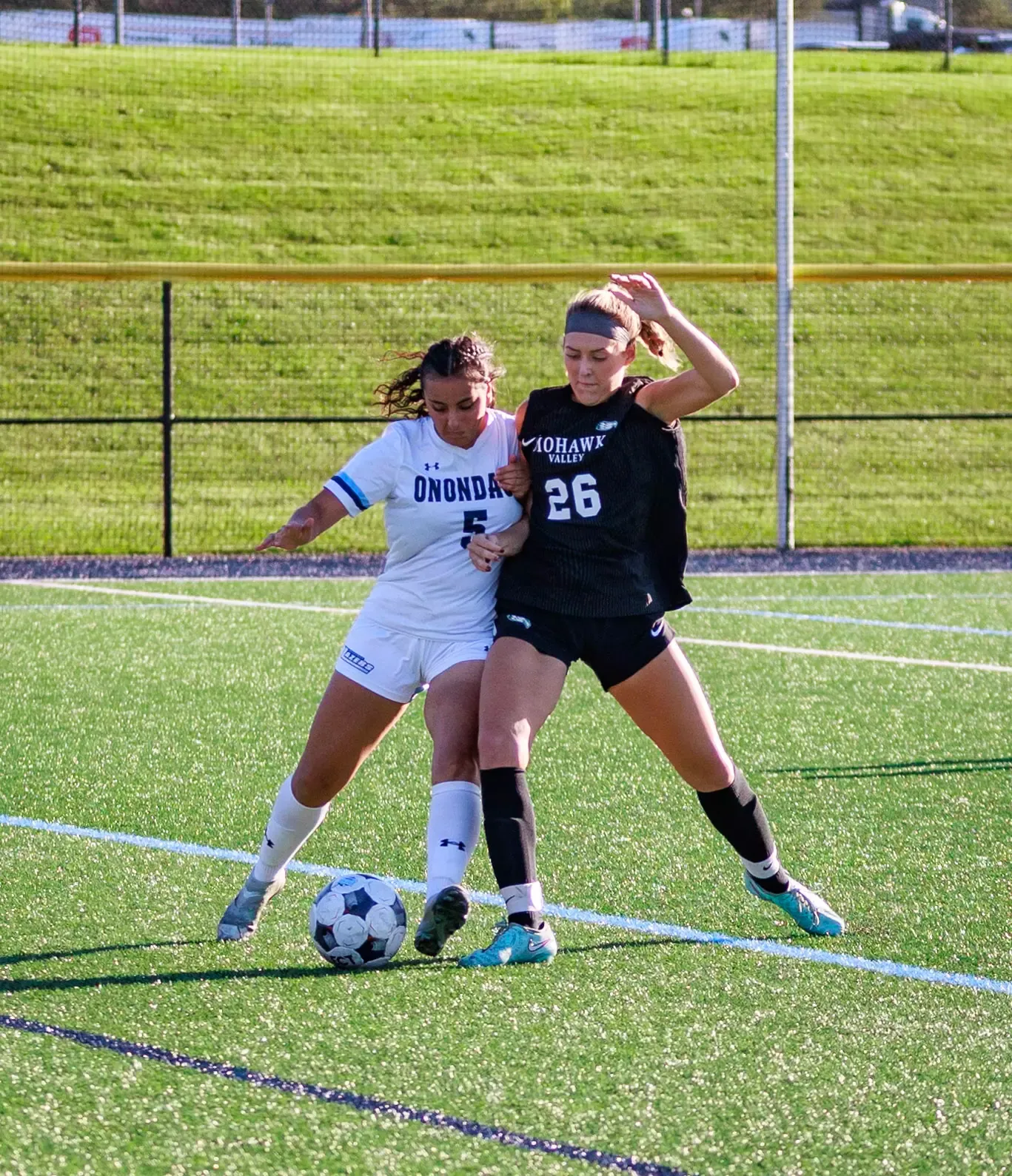 Two female soccer players compete for possession of the ball on a turf field. One wears a white Onondaga jersey with number 5, while the other wears a black Mohawk Valley jersey with number 26. Both are moving toward the ball, with the black-clad player raising one arm for balance. A fenced grassy area and sports facilities are visible in the background.