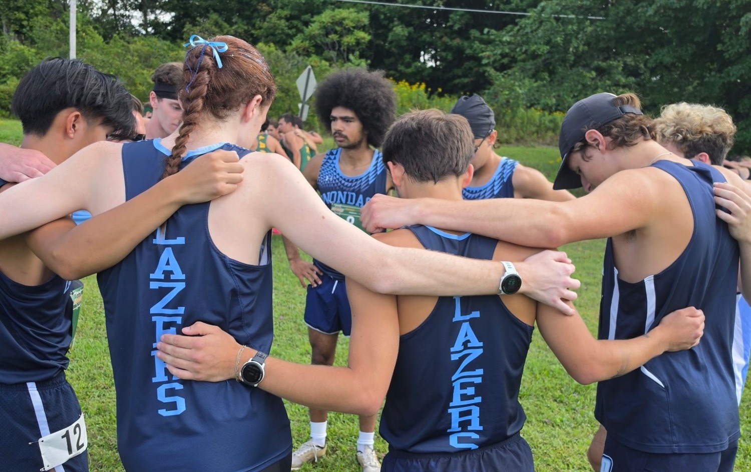 A group of teenage cross-country runners in navy uniforms stands in a close huddle on a grassy field, with their arms around one another. Several runners wear race bibs and watches, while teammates in green uniforms and wooded scenery are visible in the background.