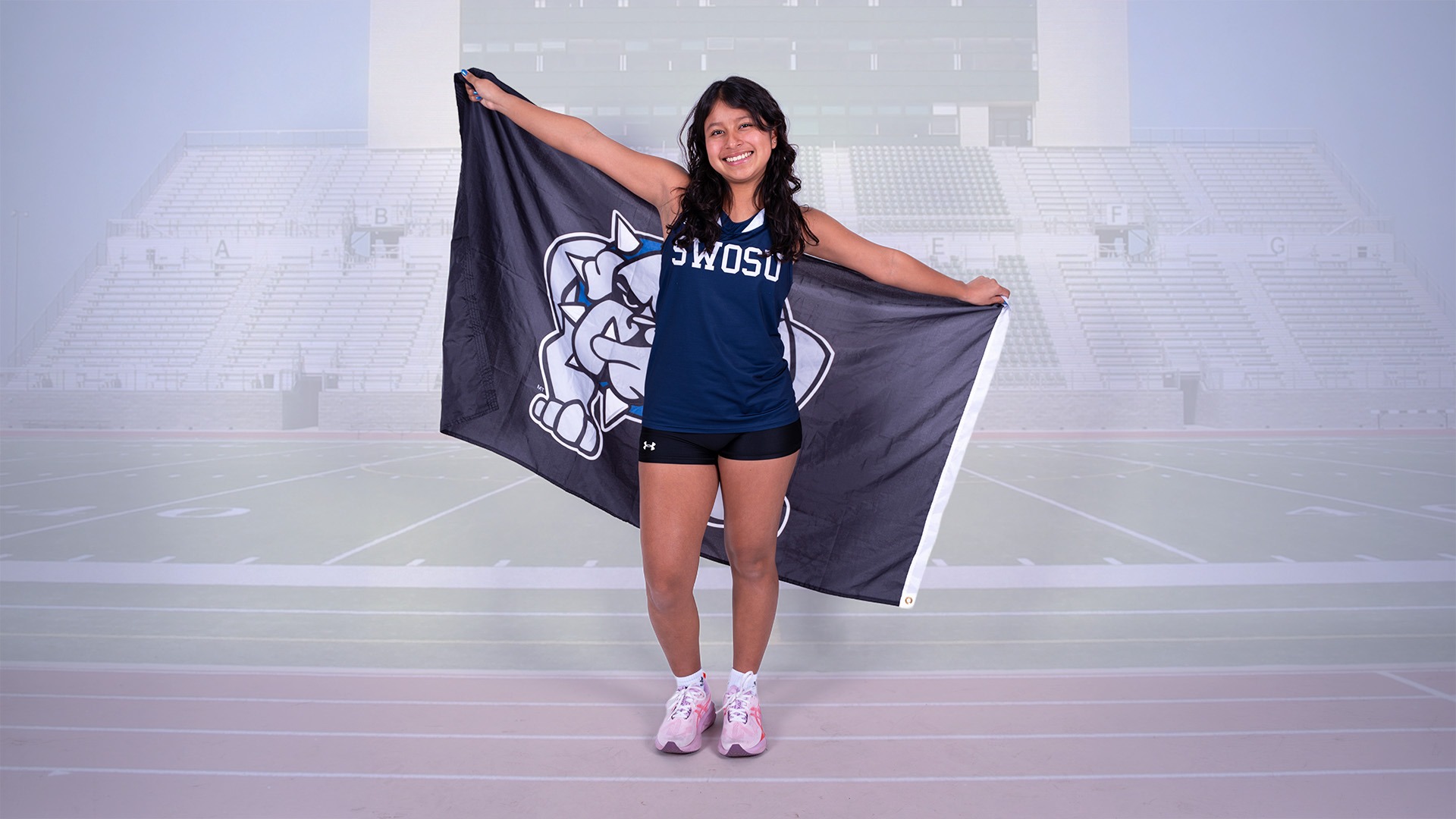 stephanie gallardo poses with a swosu flag, in the background, a very dim outline of the Red Raven Track 