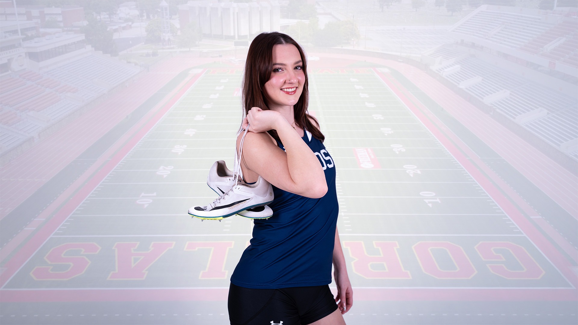 Emerson Sours poses with track shoes.  With light opacity, behind her is a photo of the Pitt State football stadium where she competed this past weekend