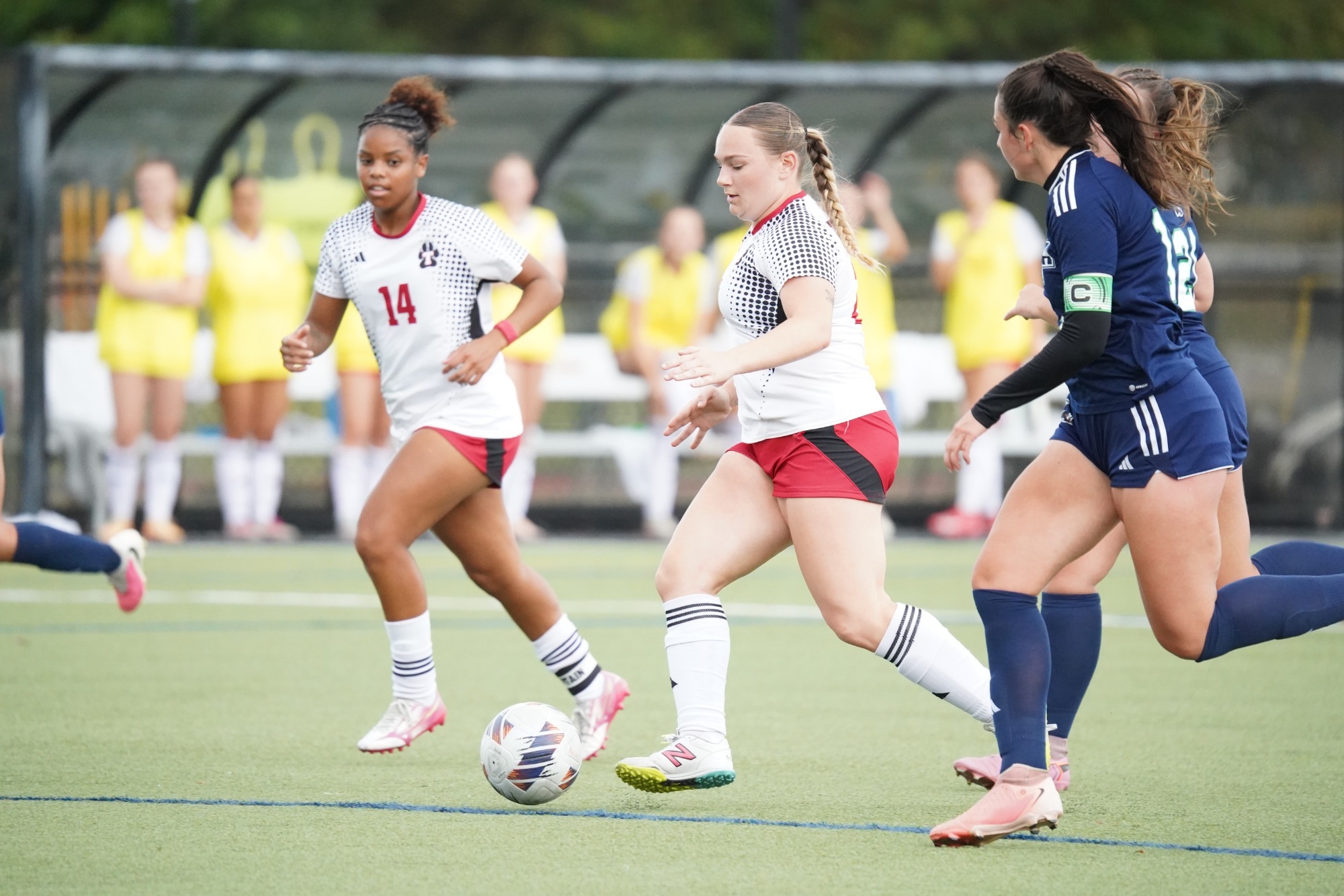 Two soccer players in white jerseys and red shorts move the ball up the field while two opposing players in navy uniforms defend. Players and team personnel stand along the sideline in the background.