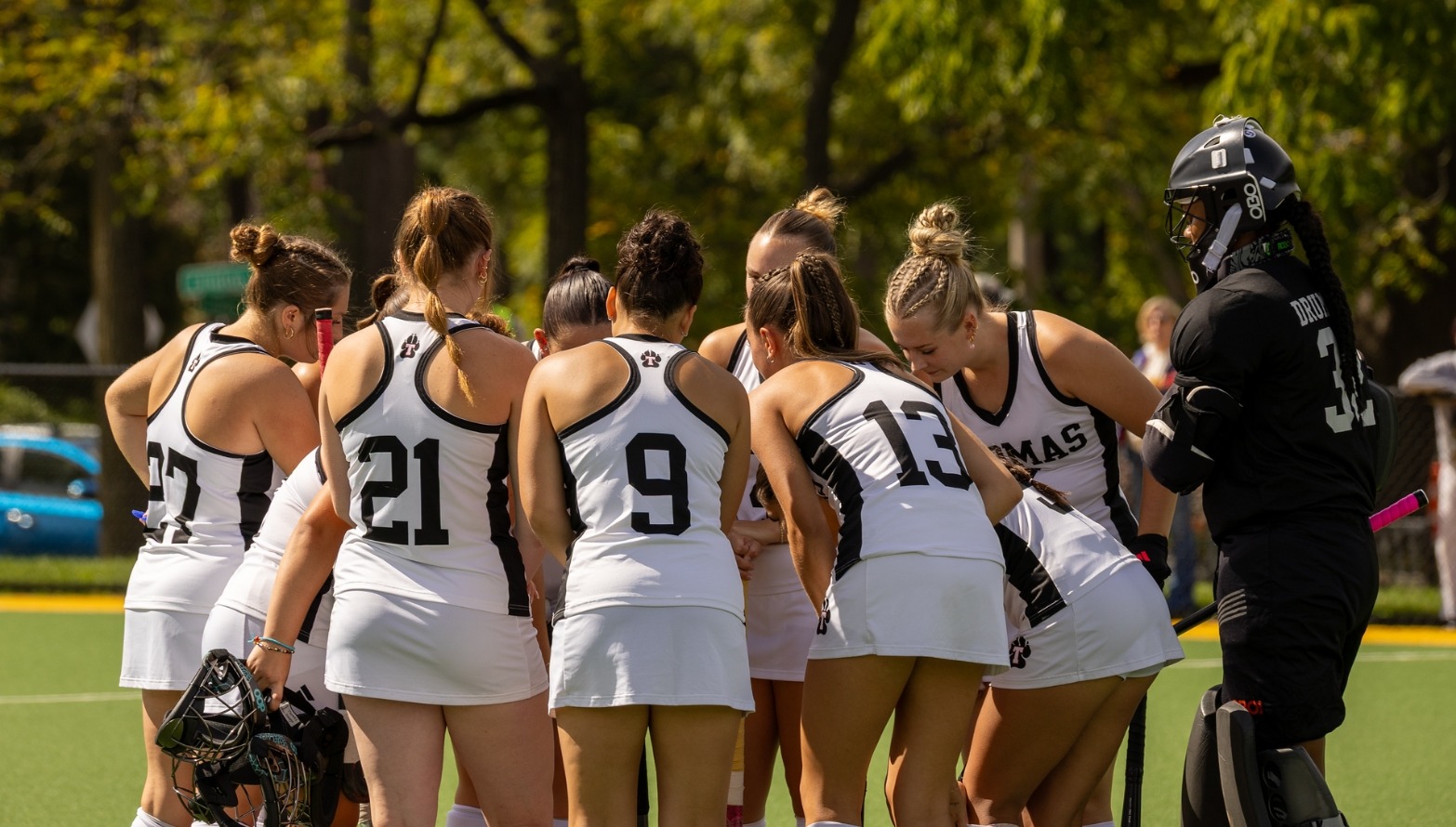 Thomas College field hockey players gather in a close huddle on the turf before play. Athletes wearing white uniforms with black trim stand shoulder-to-shoulder with sticks in hand, while a goalkeeper in full black protective gear stands beside the group. Jersey numbers including 27, 21, 9, and 13 are visible as the team leans inward near midfield. Trees and greenery in the background frame the sunny outdoor setting.