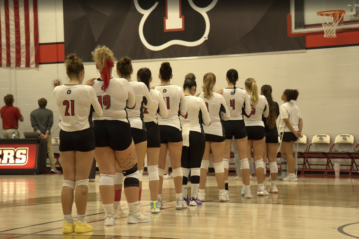 Members of the Thomas College volleyball team stand in a line on the court during the national anthem before a match. The athletes wear white jerseys with black shorts and face an American flag and scorer's table. A large Thomas College logo hangs on the gym wall above the court, with the basketball hoop and team bench visible in the background.