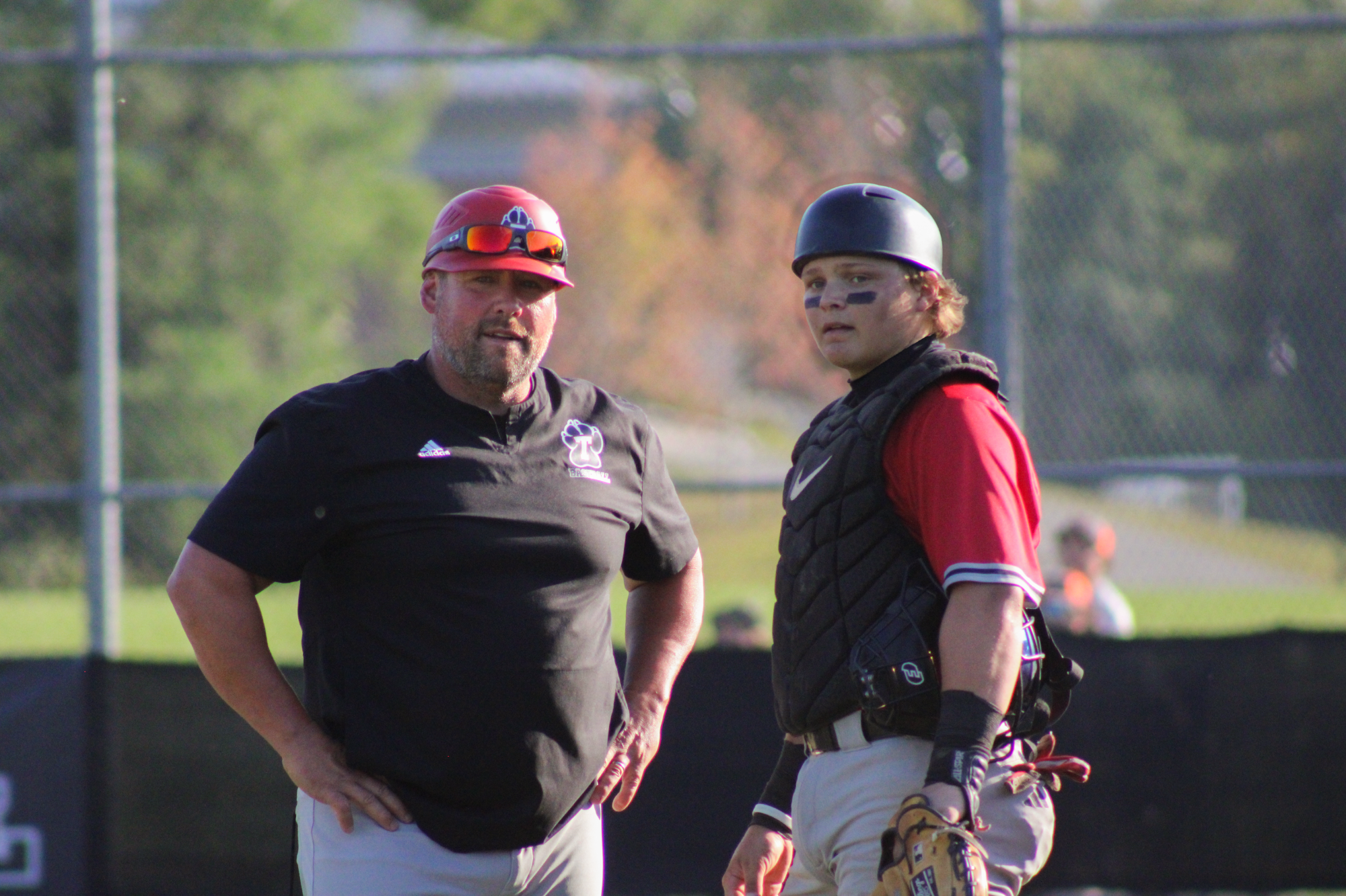 Thomas College Baseball Head Coach Rob Dippner stands on the field speaking with catcher Robby Neese during a game. Coach Dippner, wearing a black Thomas Baseball shirt and red cap with sunglasses perched on top, stands with his hands on his hips. Beside him, Neese is dressed in full catching gear, including a black chest protector, helmet, and eye black, while holding his catcher’s mitt. Both are focused on the action on the field, with the baseball diamond and outfield fence blurred in the background. The image captures a moment of communication and leadership between coach and player during competition.