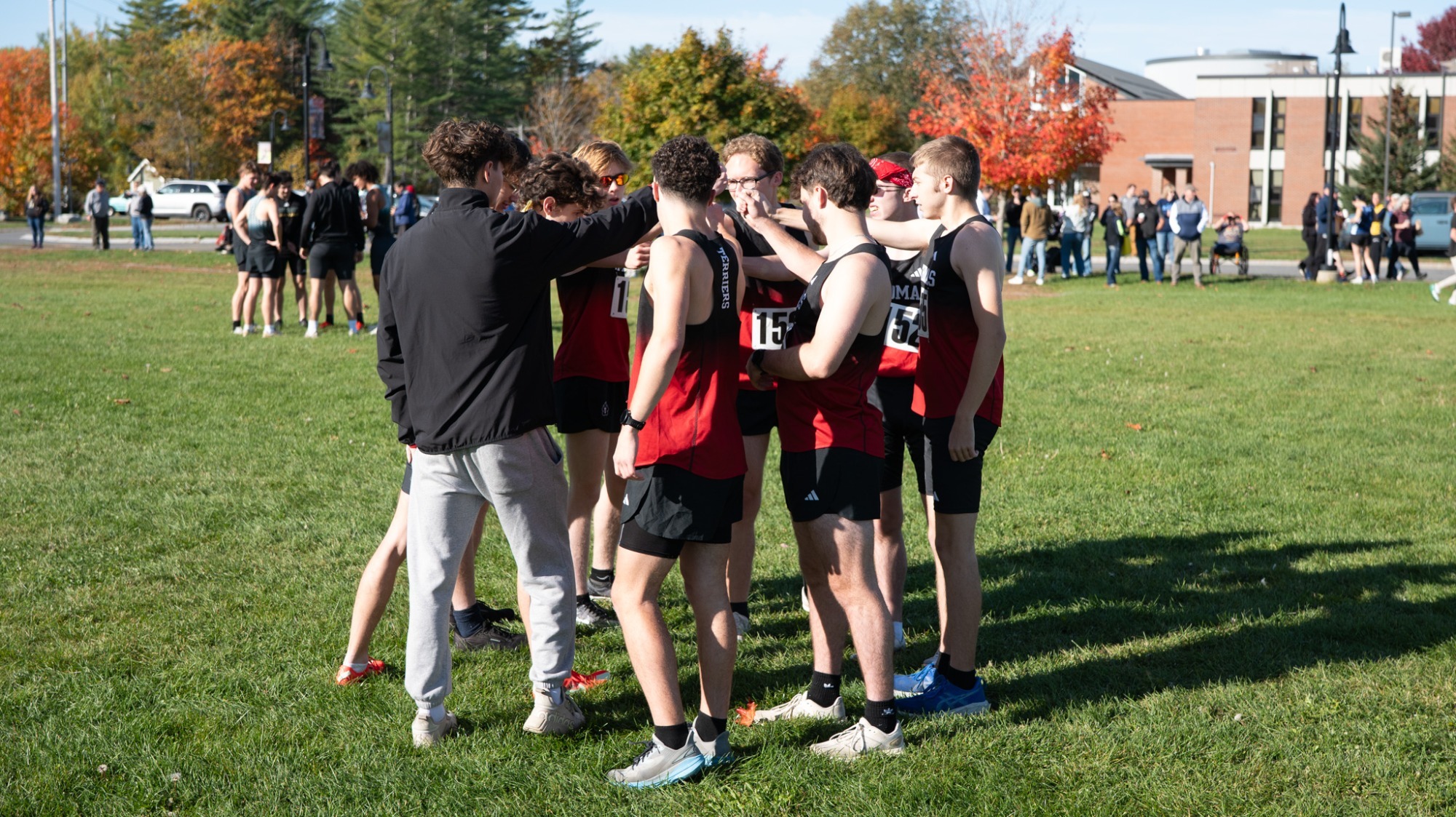 Members of the Thomas College Men's Cross Country team gather in a pre-race huddle on a grassy course with their hands together before the start of a meet. The student-athletes wear black and red Thomas College uniforms while coaches and teammates stand alongside them. Spectators, other teams, colorful fall trees, and campus buildings fill the background on a sunny autumn day.
