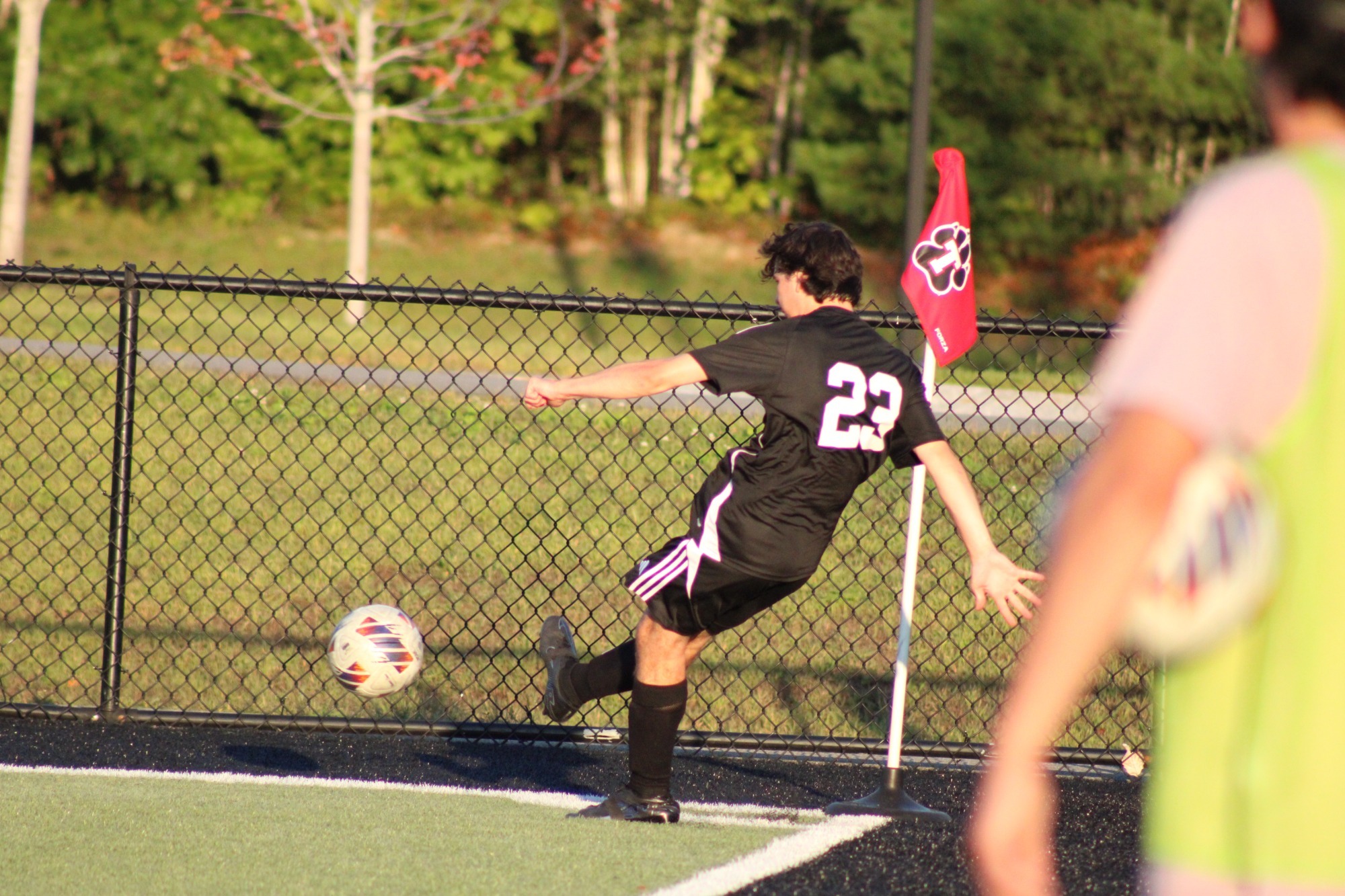 Soccer player wearing a black uniform with the number 23 kicks a ball near the corner flag on an outdoor field. A red corner flag stands beside the player, while a blurred player in a bright training vest appears in the foreground. A chain-link fence and green trees are visible in the background under warm sunlight.