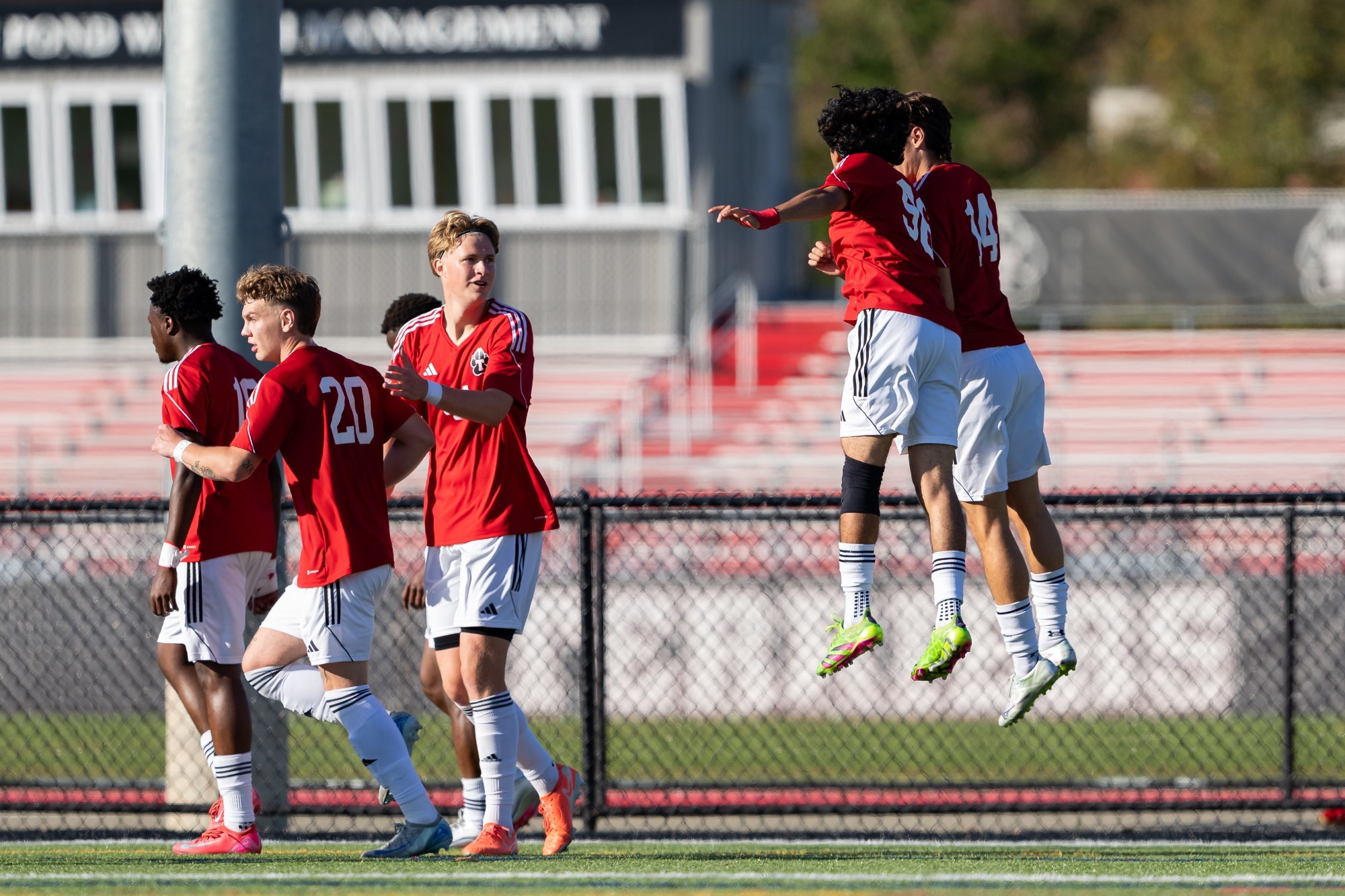 Several soccer players in red jerseys and white shorts gather on the field during a match. Two players leap into the air in celebration near the right side of the image, while teammates walk nearby. A black chain-link fence separates the field from bleachers and a building in the background. The scene is brightly lit by afternoon sunlight.