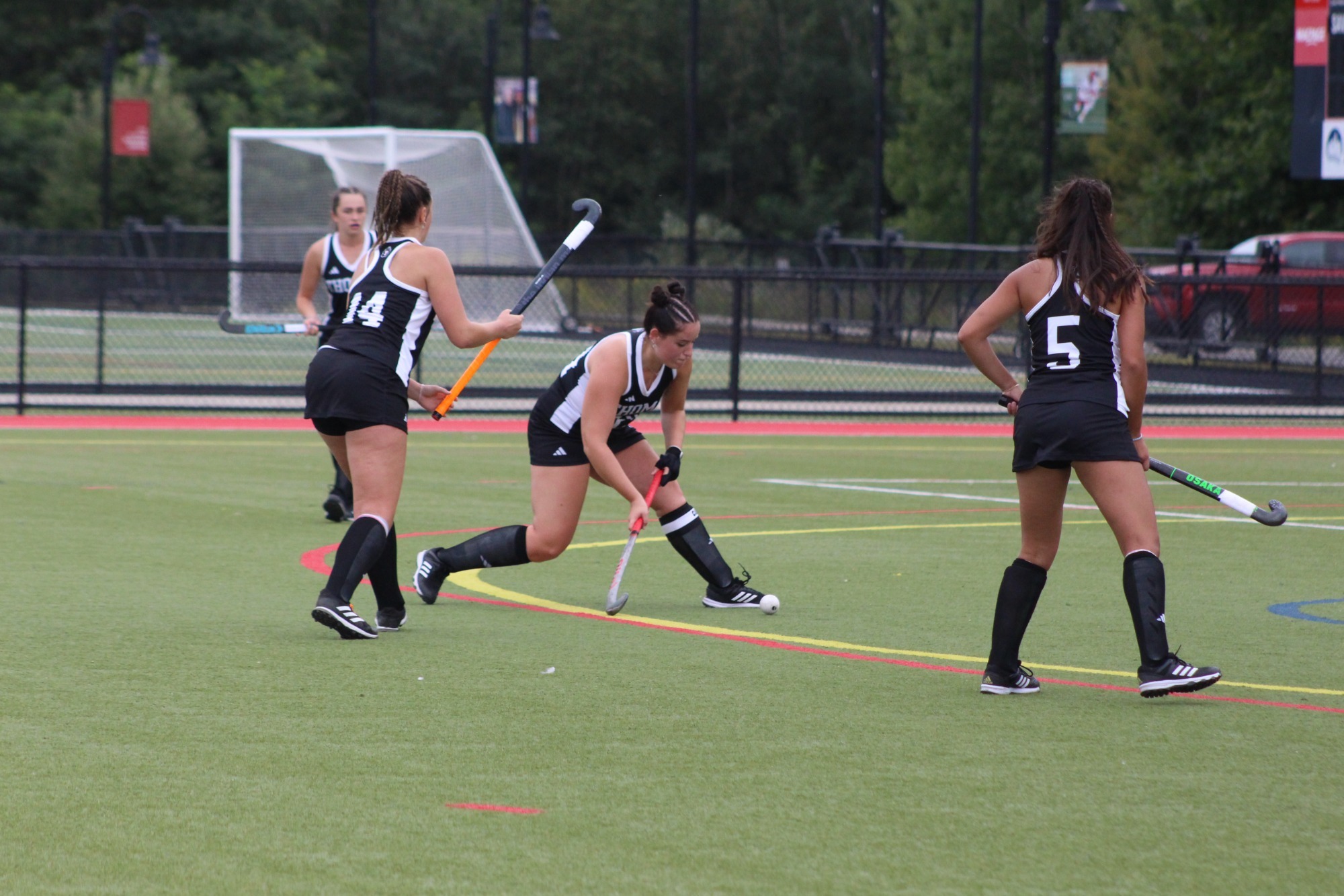 Three field hockey players in black uniforms move the ball up a turf field during game action. One player bends low while dribbling the ball with a stick, while teammates wearing numbers 14 and 5 position themselves nearby for support. A goal cage, fencing, and trees are visible in the background on an overcast day.
