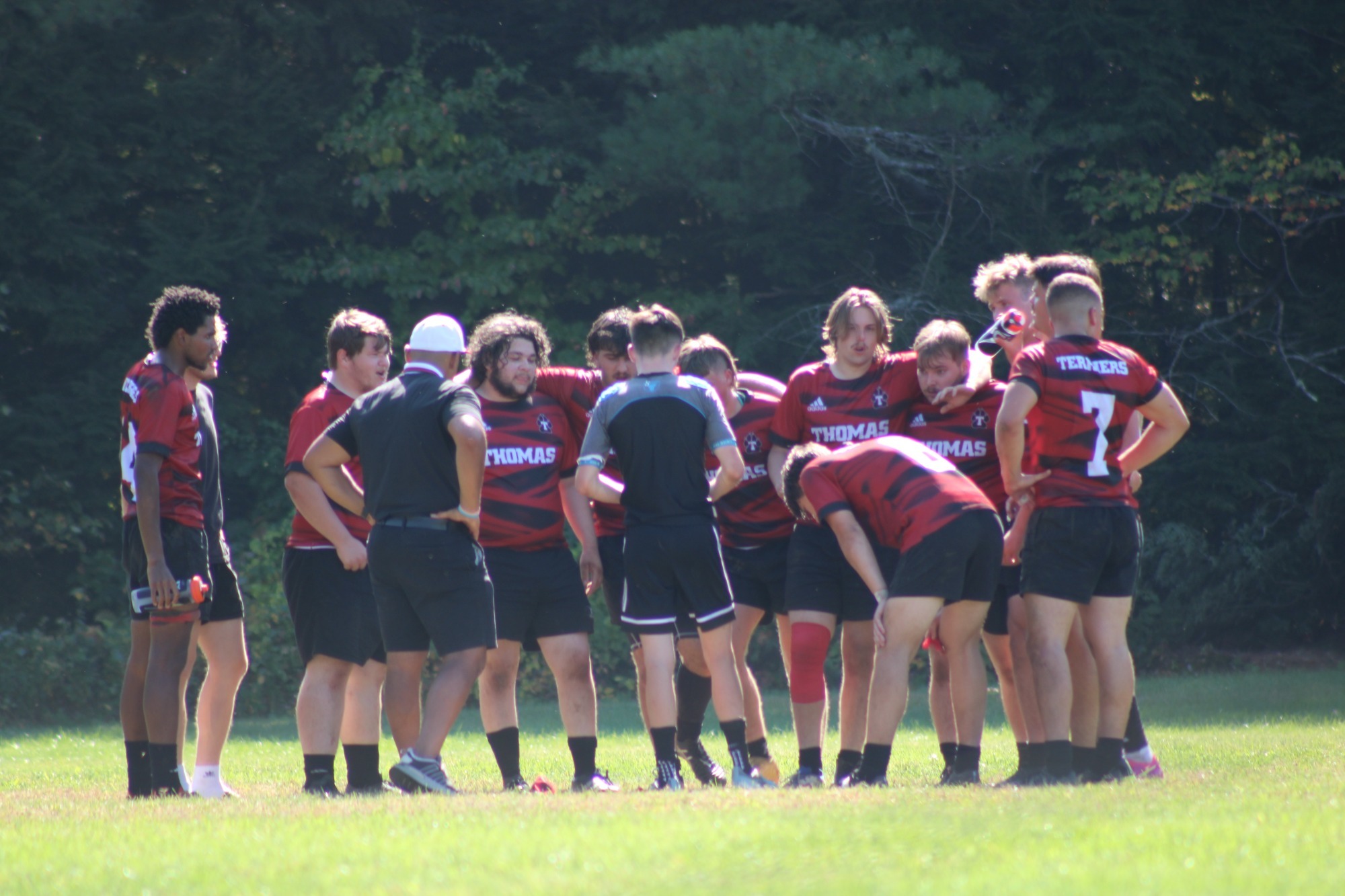 Members of a rugby team in red-and-black striped jerseys gather in a huddle on a grassy field during a match. Several players stand shoulder to shoulder around coaches and teammates as they listen to instructions, with visible jersey numbers including 7. Trees and wooded areas form the backdrop, and bright sunlight illuminates the scene.