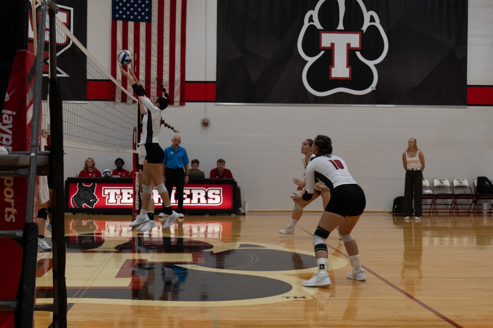 Alt text:  Volleyball action inside a college gymnasium. A player in a white jersey jumps at the net to play the ball while teammates in white jerseys prepare on the court. Large American flags and a black banner featuring a red-and-white “T” logo hang on the wall behind the court, and a scorer’s table displays the word “Terriers.” The polished hardwood floor reflects the players and gym lighting.