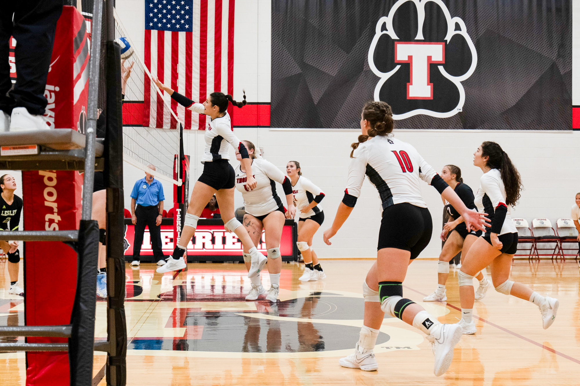 Indoor volleyball game in a gymnasium with multiple players in white jerseys and black shorts positioned on the court. One player jumps at the net with an outstretched arm to play the ball while teammates prepare behind and around the play. A referee stand and net post are visible in the foreground. An American flag and a large paw-print logo with a red “T” hang on the wall above the court. The polished wood floor reflects the players and gym lighting.