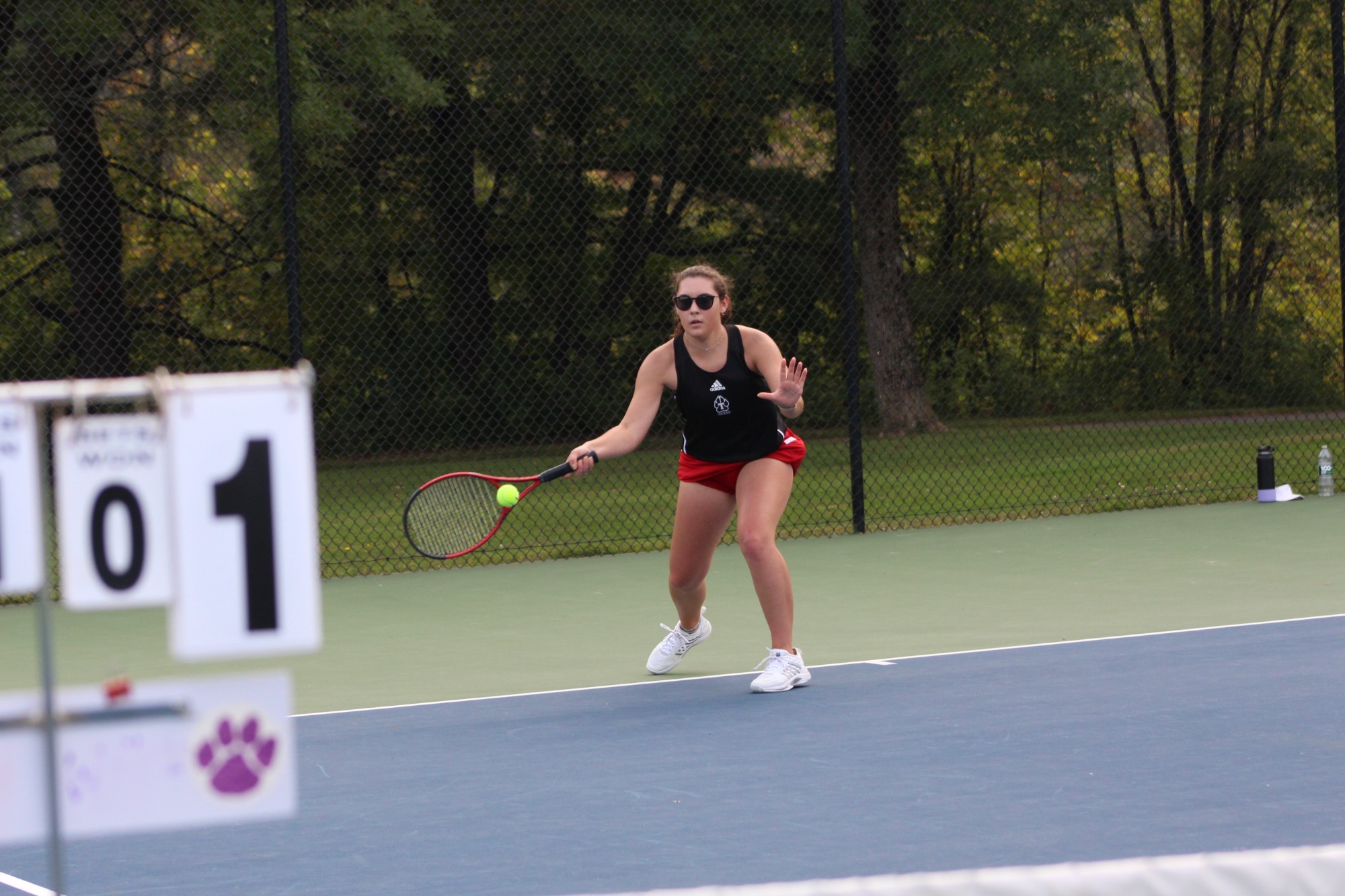 Alt text:  A college tennis player wearing a black top, red shorts, and sunglasses prepares to return a shot on an outdoor tennis court. Holding a racket in front of the body, the player tracks a bright yellow tennis ball approaching near the baseline. A courtside scoreboard displaying the number 