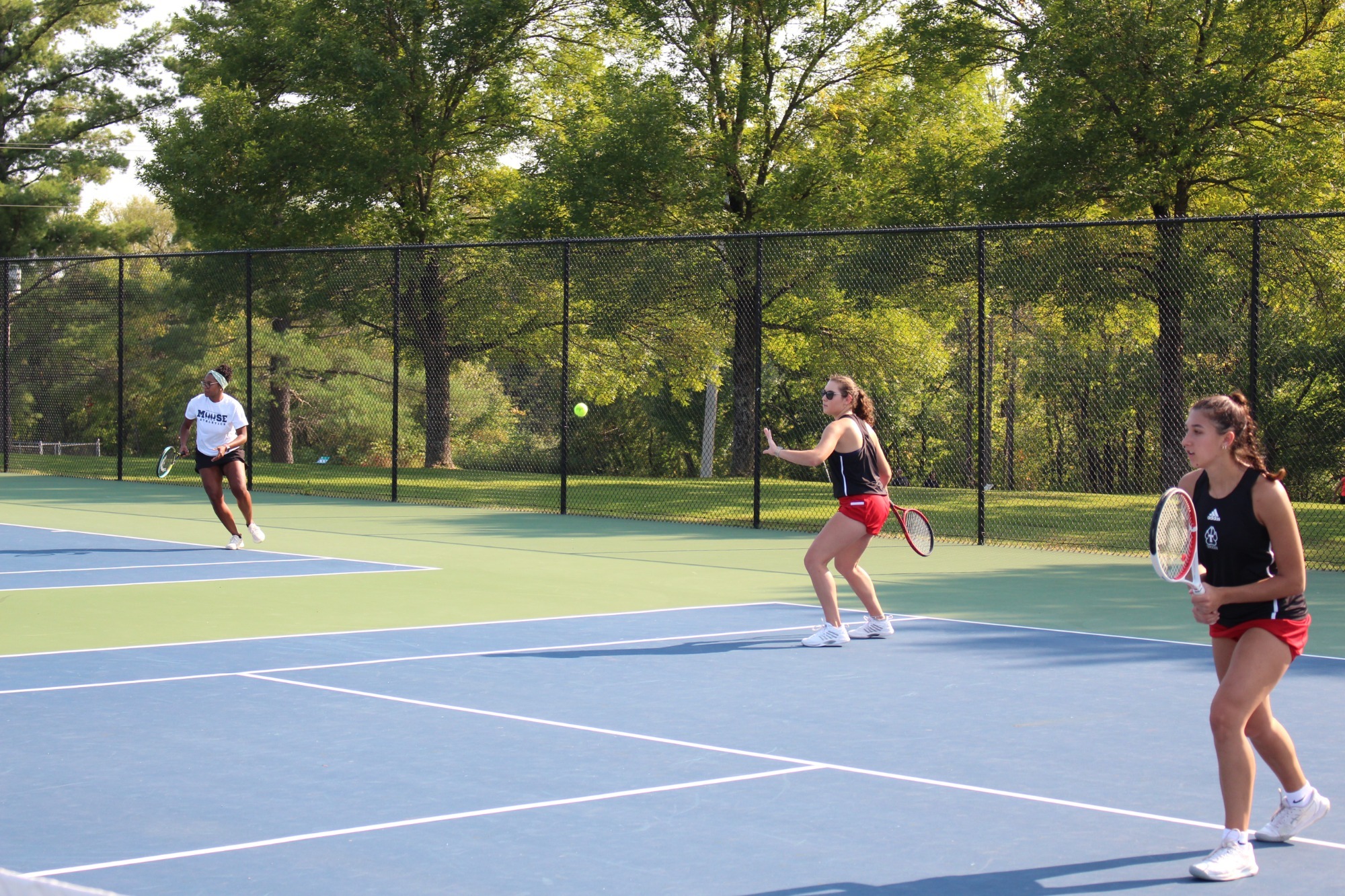 Two college tennis players in black tops and red shorts prepare for doubles play on an outdoor tennis court. One player near the net positions a racket to return an approaching ball, while a teammate waits in a ready stance closer to the camera. An opposing player is visible on the far side of the court. The match takes place on a sunny day, with a chain-link fence and tall green trees surrounding the court.