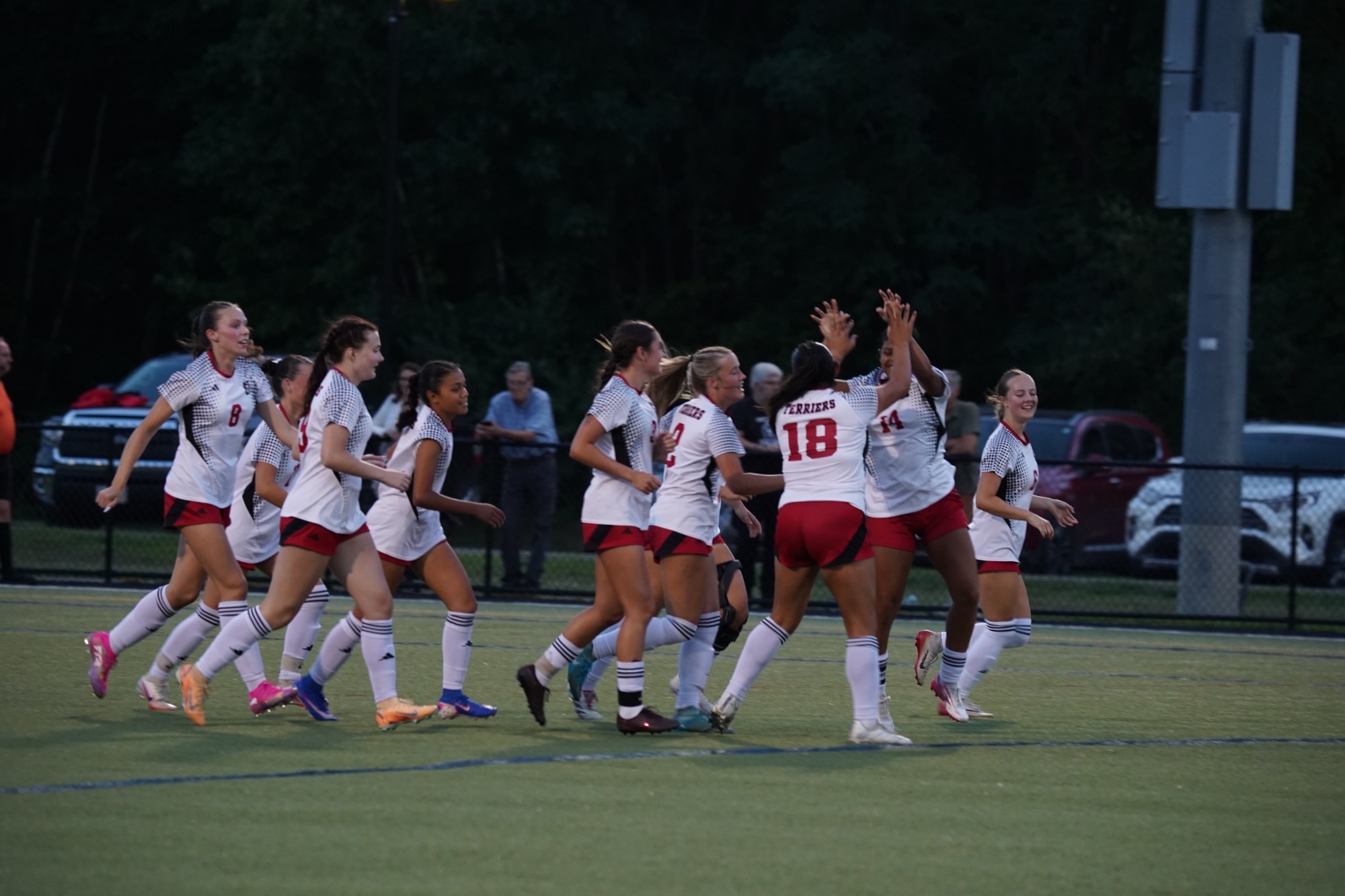A group of soccer players in white jerseys with red accents and red shorts gather on a turf field during an evening match. Several players are raising their hands in celebration near midfield while teammates run toward them. Spectators stand behind a fence in the background, with parked vehicles and a wooded area beyond the field. The scene is lit by stadium lights against a darkening sky.