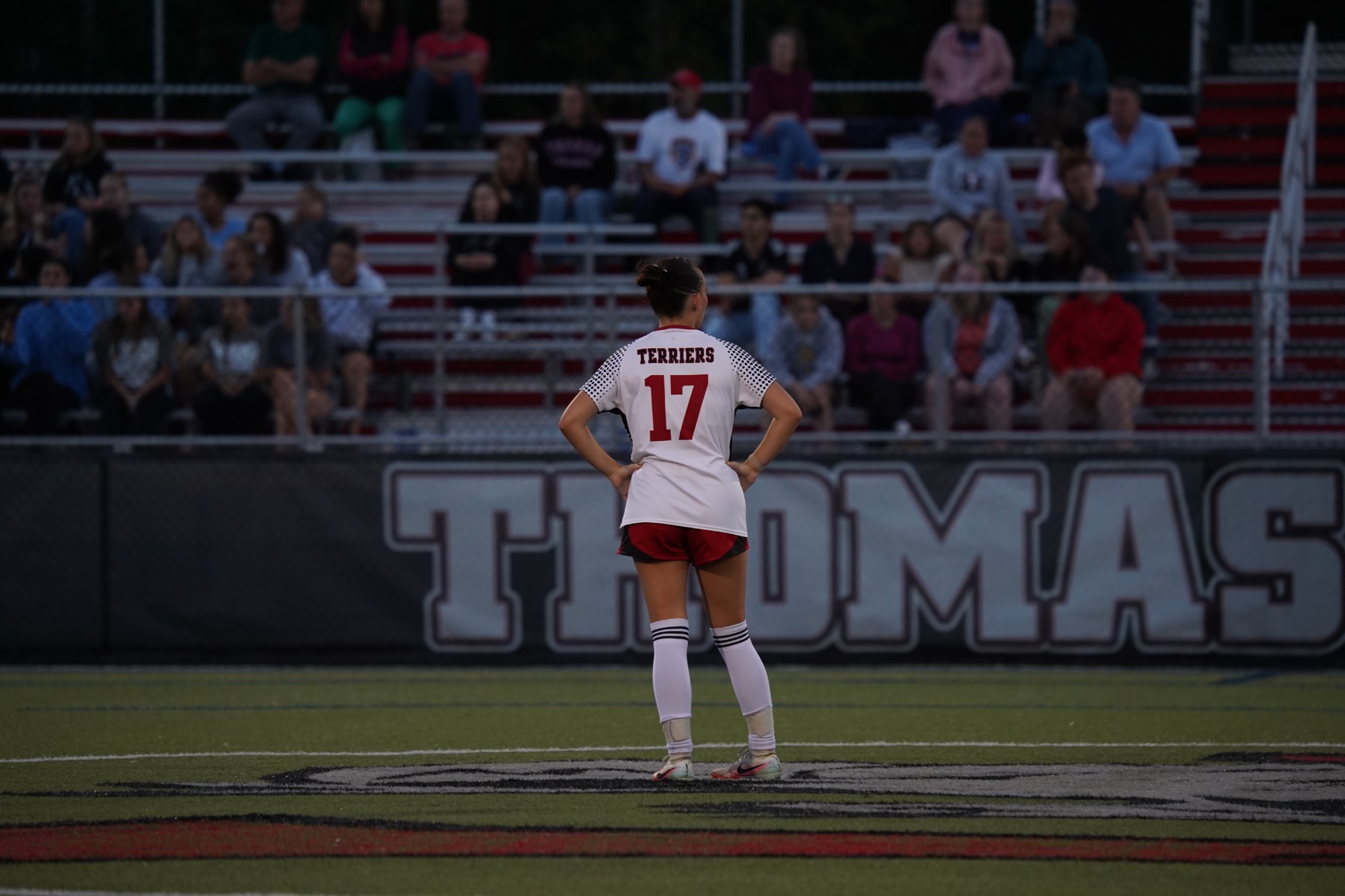 A soccer player wearing a white jersey with red shorts and the number 17 stands at midfield on a turf field with hands on hips. The back of the jersey reads “TERRIERS.” In the background, spectators sit in red-and-white bleachers behind a fence. Large lettering spelling “THOMAS” is visible on a wall behind the player. The scene takes place during an evening match under stadium lighting.