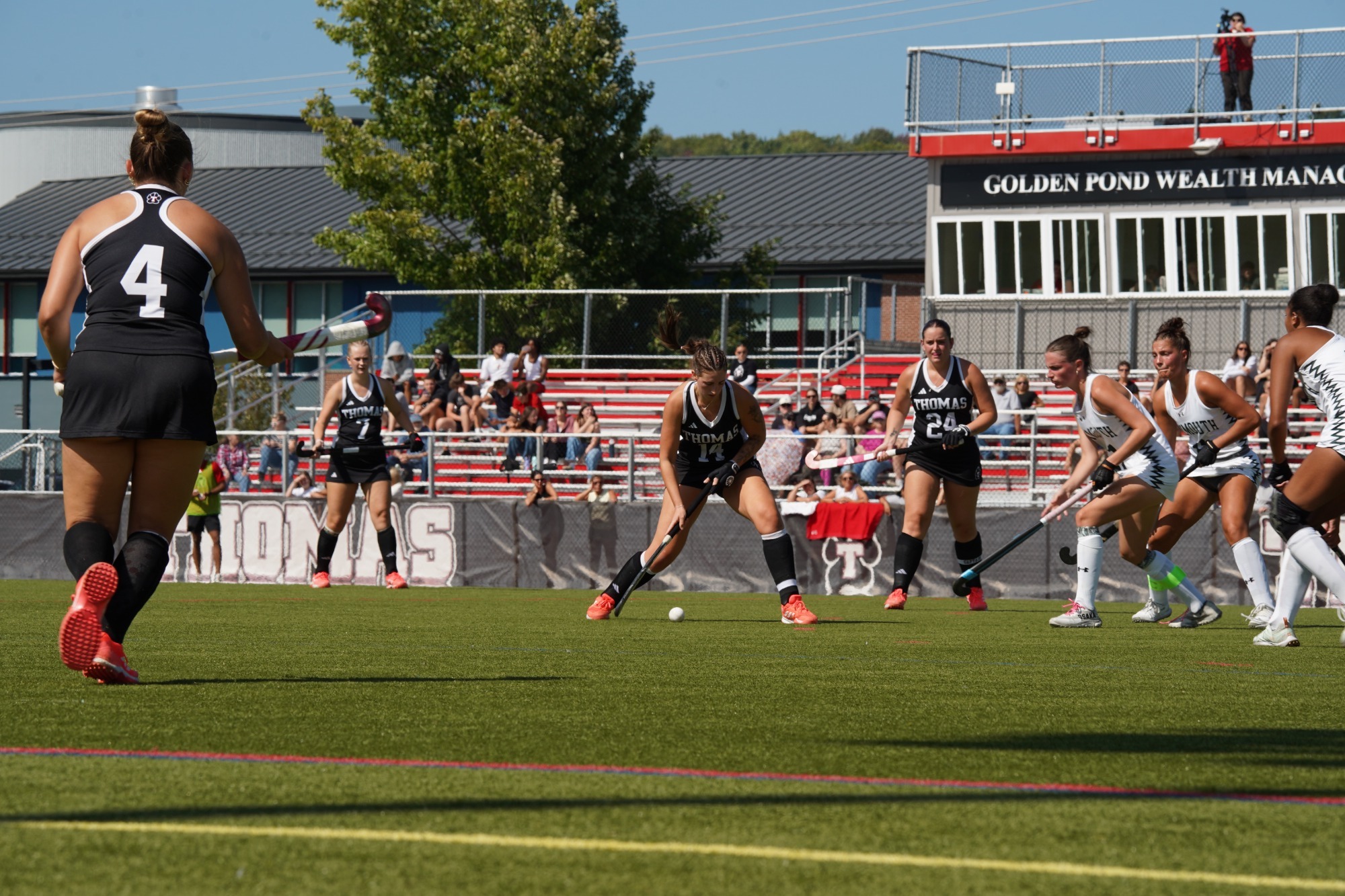 Thomas College field hockey players compete for possession of the ball during a home game on a sunny afternoon. A player wearing number 14 in a black Thomas uniform controls the ball while teammates move into position nearby. Opposing players in white uniforms close in defensively. Spectators sit in the red bleachers behind the field, with the press box and athletic facilities visible in the background.
