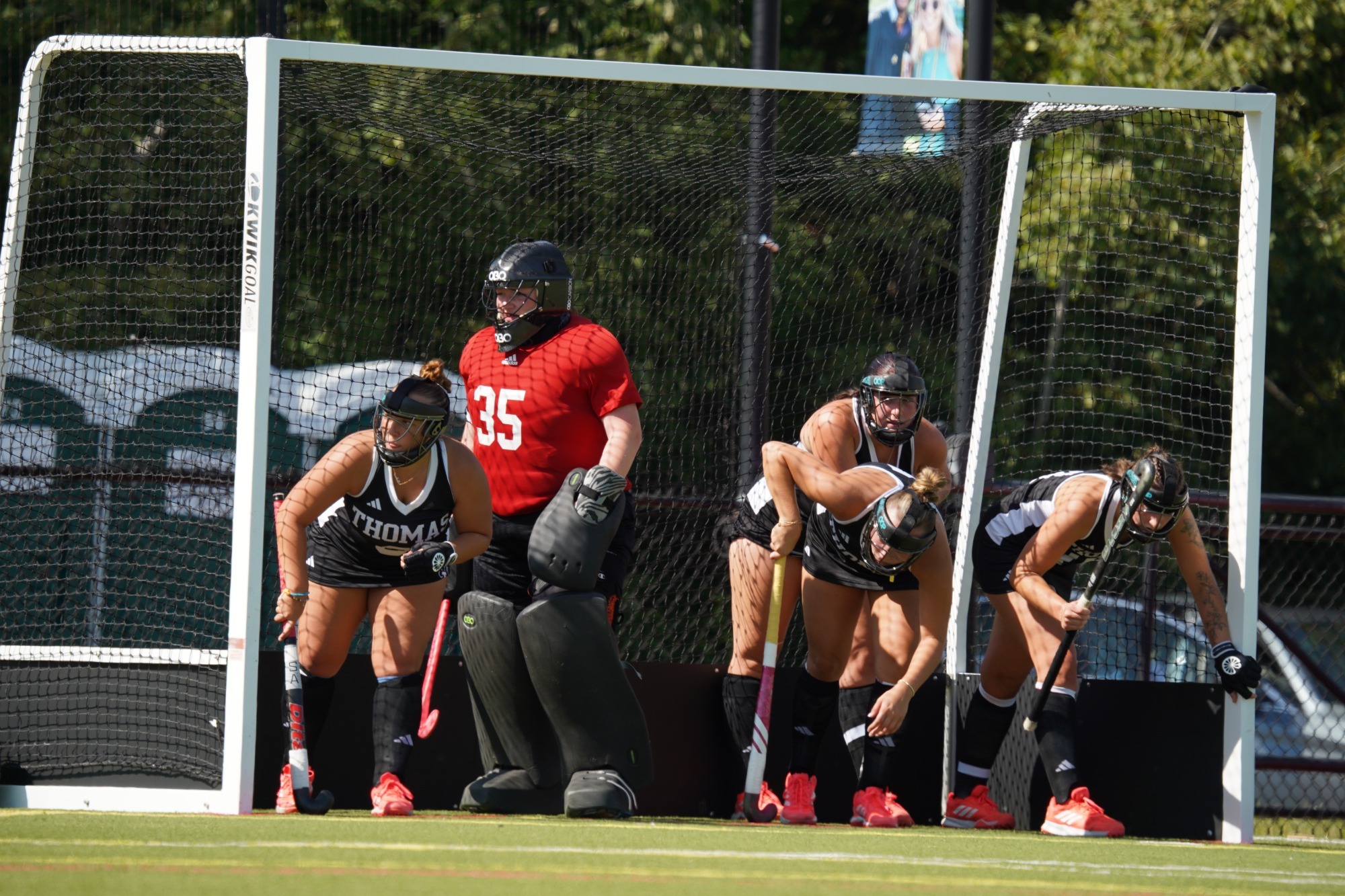 Thomas College field hockey players line up inside the goal cage to defend a penalty corner during a home game. A goalkeeper wearing red protective gear stands ready in front of the net while four defenders in black Thomas College uniforms crouch in position along the goal line, preparing to sprint out as play begins. The scene takes place on a sunny afternoon with trees and athletic facilities visible beyond the field.