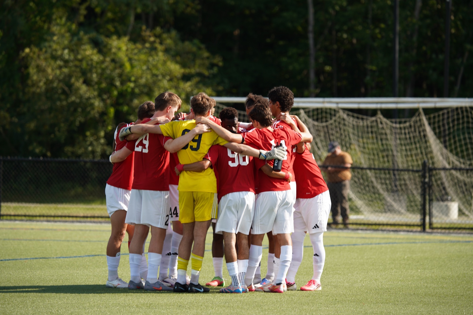Members of the Thomas College men’s soccer team stand in a tight huddle on the field before competition, with arms around each other near midfield. Players wear red jerseys and white shorts, while the goalkeeper stands in the center in a bright yellow uniform. A soccer goal, fencing, and trees are visible in the background during a sunny afternoon at the outdoor stadium.