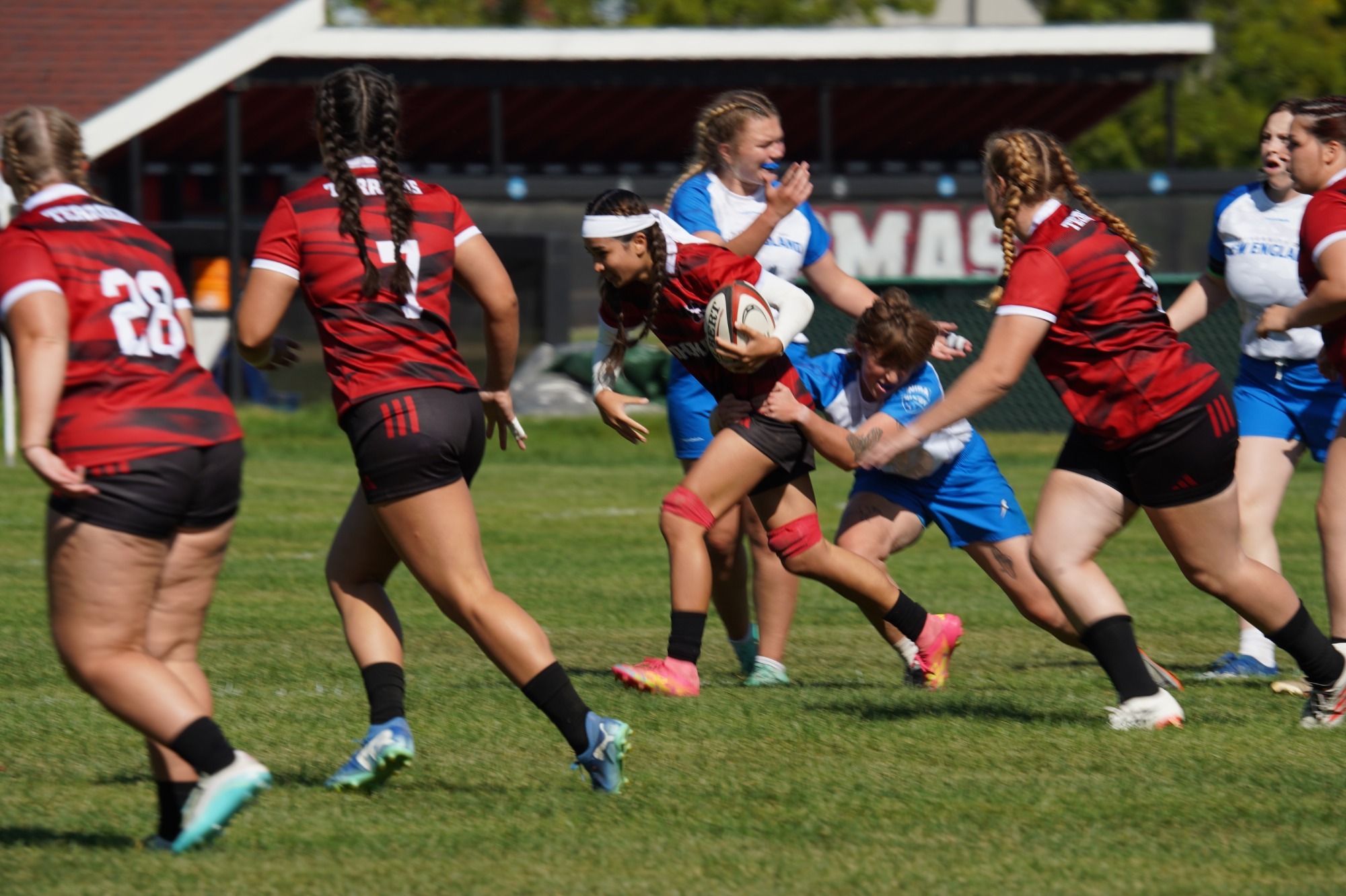 A Thomas College rugby player carries the ball upfield while being tackled by an opposing player during a match on a sunny afternoon. Several teammates in red-and-black striped jerseys run alongside in support, while additional defenders in blue-and-white uniforms close in on the play. The action takes place on a grass field with athletic facilities visible in the background.