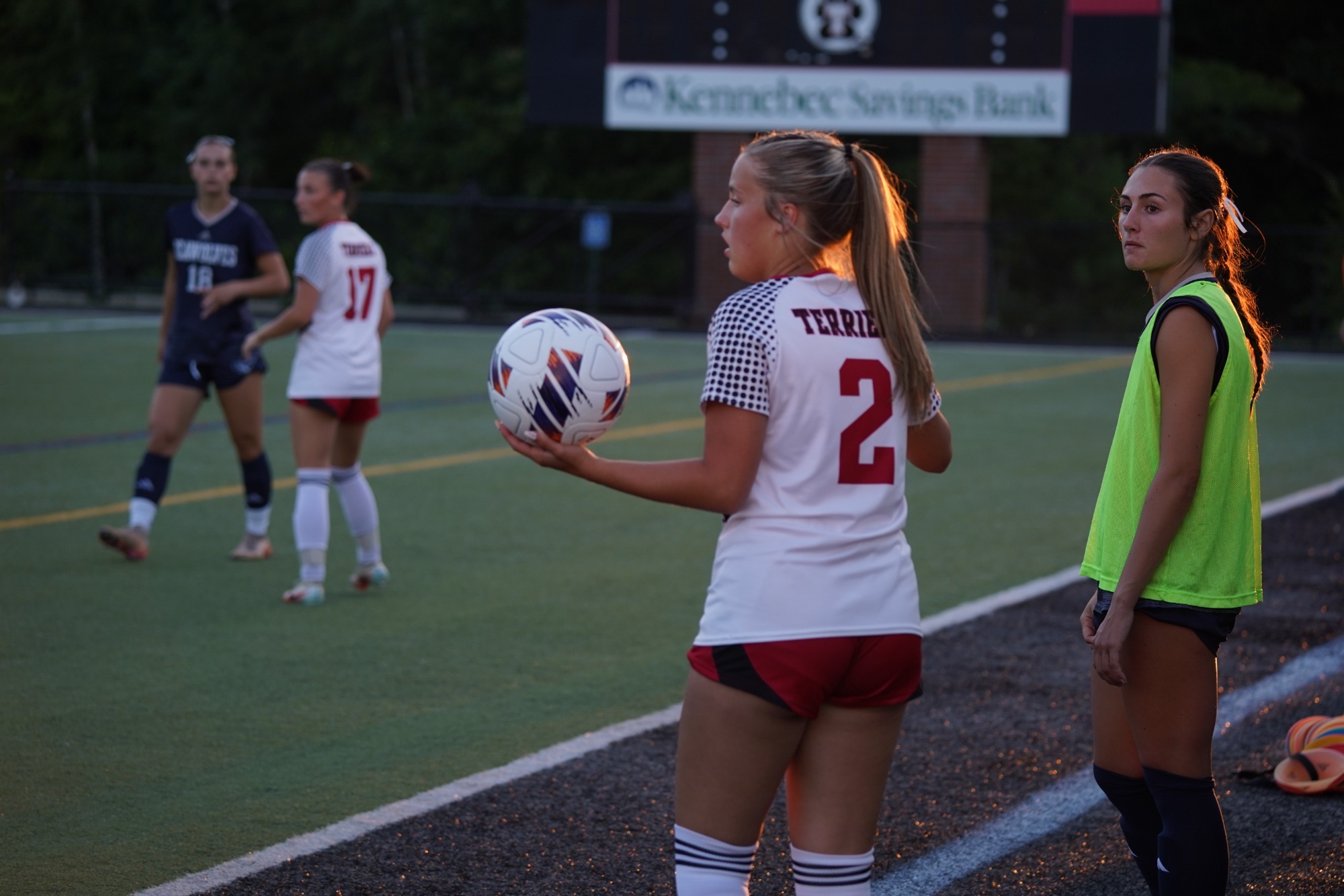 A Thomas College women’s soccer player wearing jersey number 2 stands at the sideline holding a soccer ball, preparing for a throw-in during an evening match. Another Thomas College player wearing a training pinnie waits nearby, while teammates and opposing players are positioned on the field in the background. Stadium lights and a scoreboard sponsored by Kennebec Savings Bank are visible behind the action as the sun sets over the field.