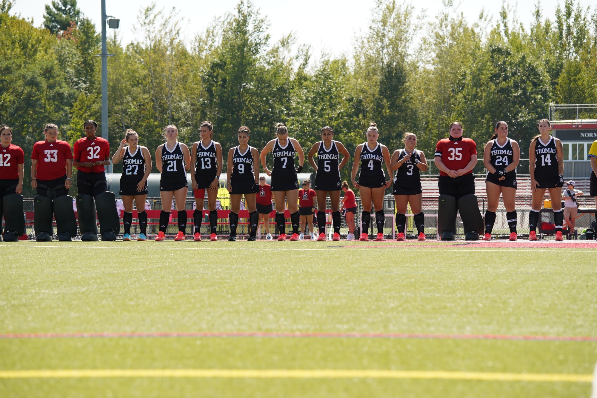 Members of the Thomas College field hockey team stand in a line along the sideline before a game on a sunny day. Players wear black uniforms with white lettering and numbers, while goalkeepers are dressed in red jerseys and protective leg pads. The team is positioned on a turf field with bleachers, trees, and athletic facilities visible in the background.