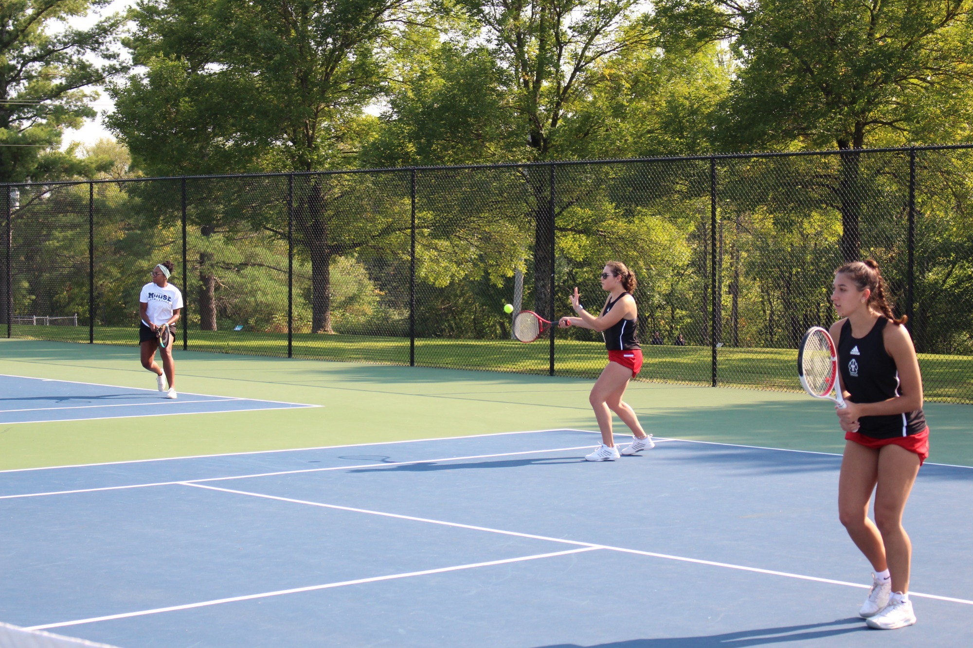 Two Thomas College women’s tennis players in black tops and red shorts prepare for a doubles point on an outdoor court. One player at the net readies a forehand volley as the tennis ball approaches, while a teammate stands near the baseline holding a racket in a ready position. An opposing player in a white shirt waits across the net, with a chain-link fence and tall green trees surrounding the court on a sunny afternoon.