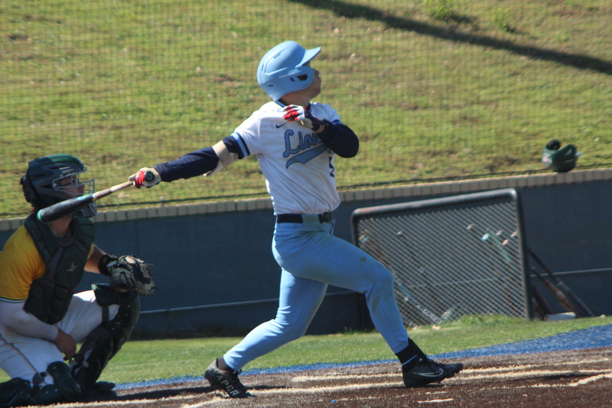 A baseball player in a blue helmet, white jersey and blue pants swings at pitch in front of the catcher. 