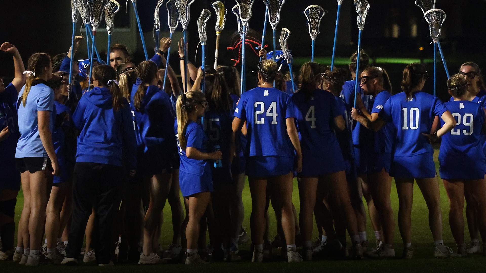 U A H women's lacrosse team huddled up with their sticks in the sky. They are wearing blue uniforms.