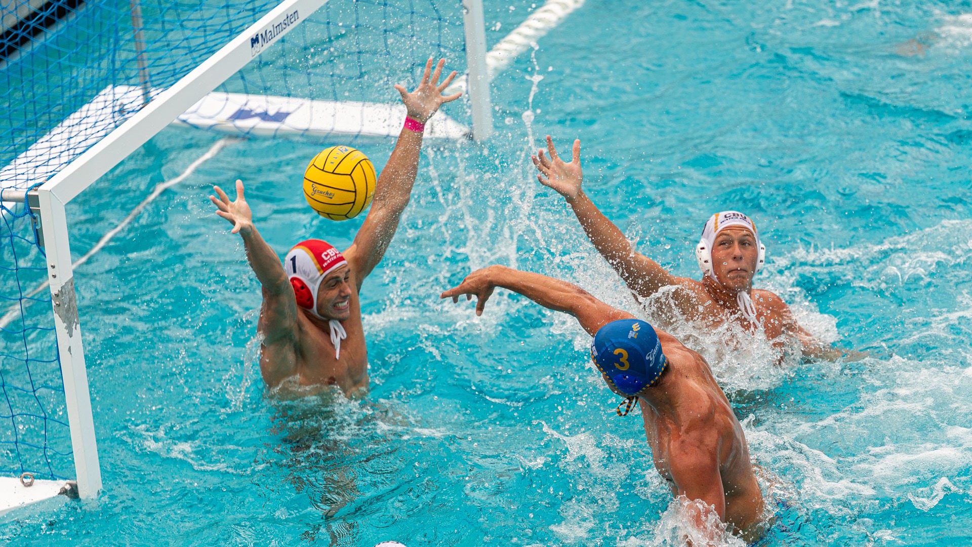 An overhead view as a Gaucho Men's Water Polo player throws a shot at the goal from very close range. The opposing goalkeeper has both hands in the air to try and block it, and a defender reaches his arm fruitlessly towards the shot from the right of the frame