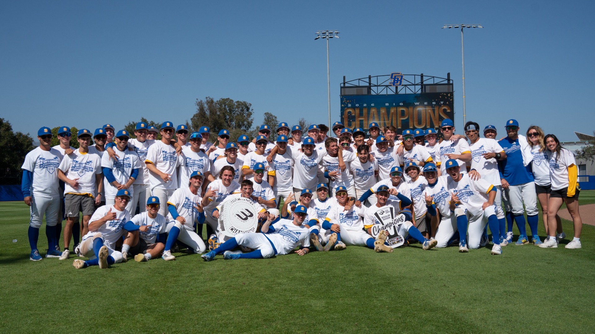The UC Santa Barbara Baseball team, all wearing white championship t-shirts over their jerseys, celebrate clinching The Big West Regular Season Championship on the field at Caesar Uyesaka Stadium. Behind the mob of baseball players, part of the Champions graphic is visible on the stadium's videoboard