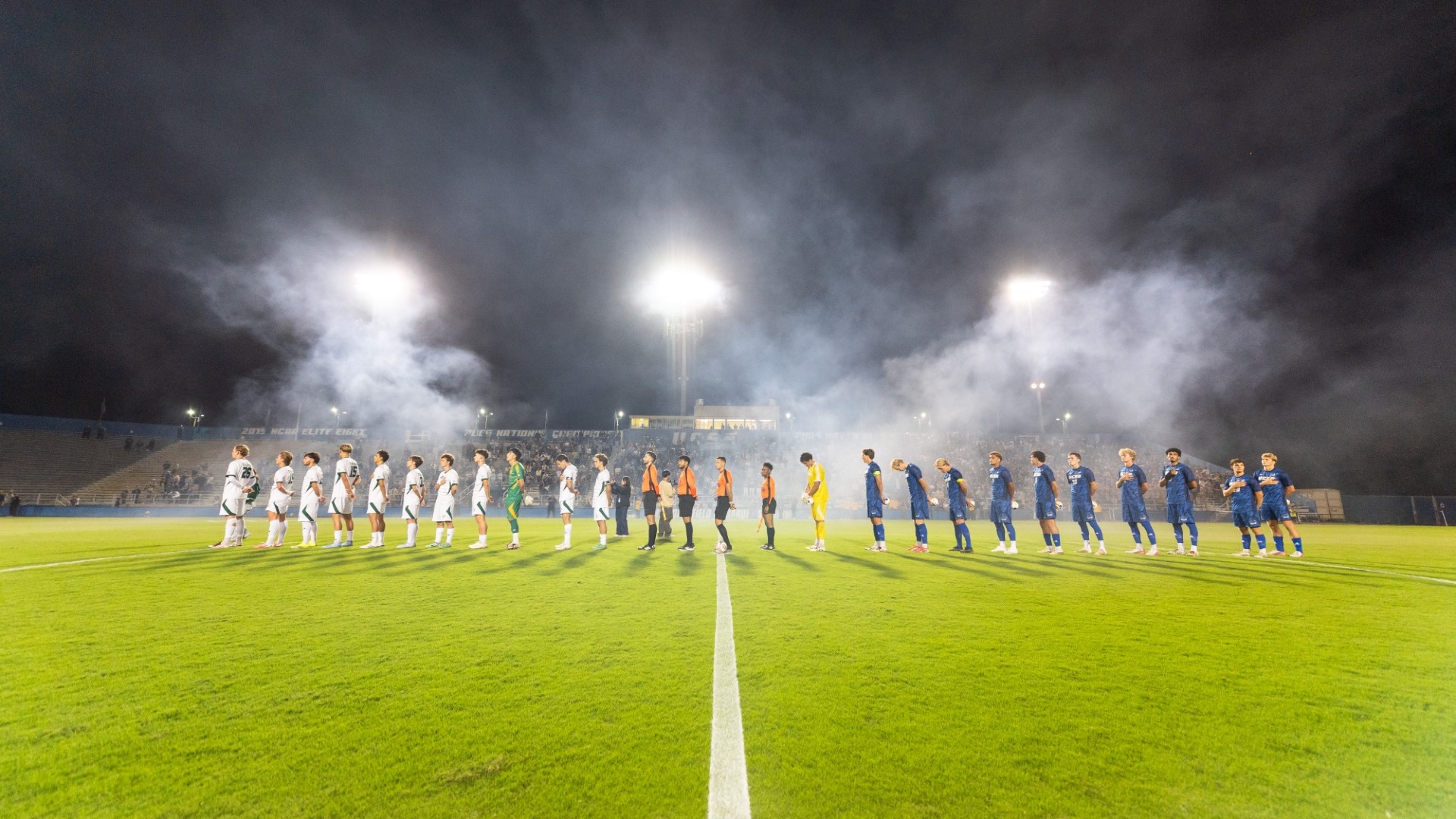 Smoke hangs in the night air at Harder Stadium as the starting elevens of Cal Poly and UC Santa Barbara Men's Soccer stand in the middle of the field for the national anthem.