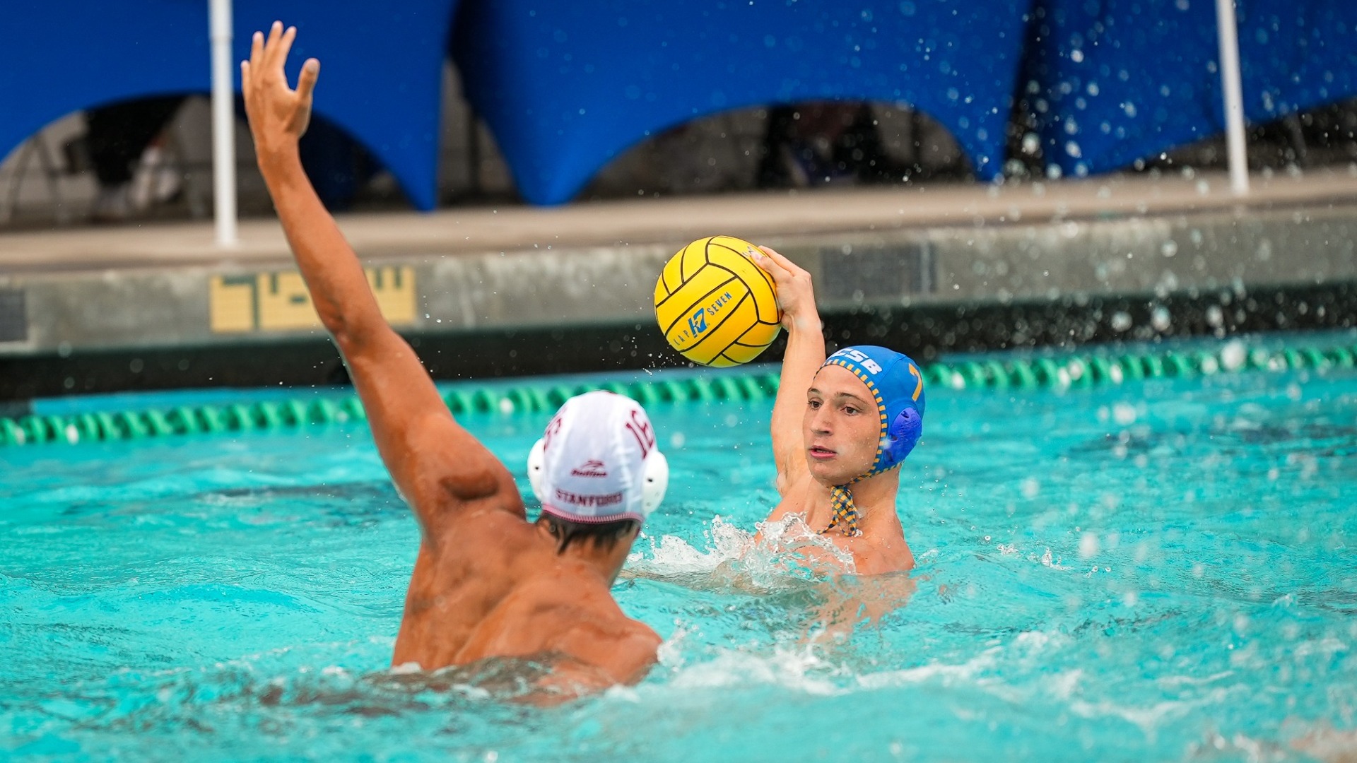 #7 Luigi Canepa lifts the ball out of the water to start winding up for a throw, while a Stanford defender raises an arm to try and block the pass.