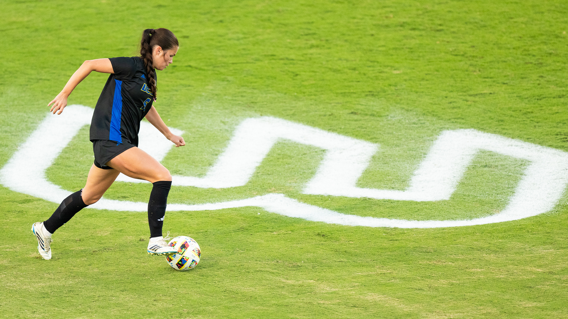 Dakota Watterson dribbles the ball across the field with the painted Big West logo behind her. 