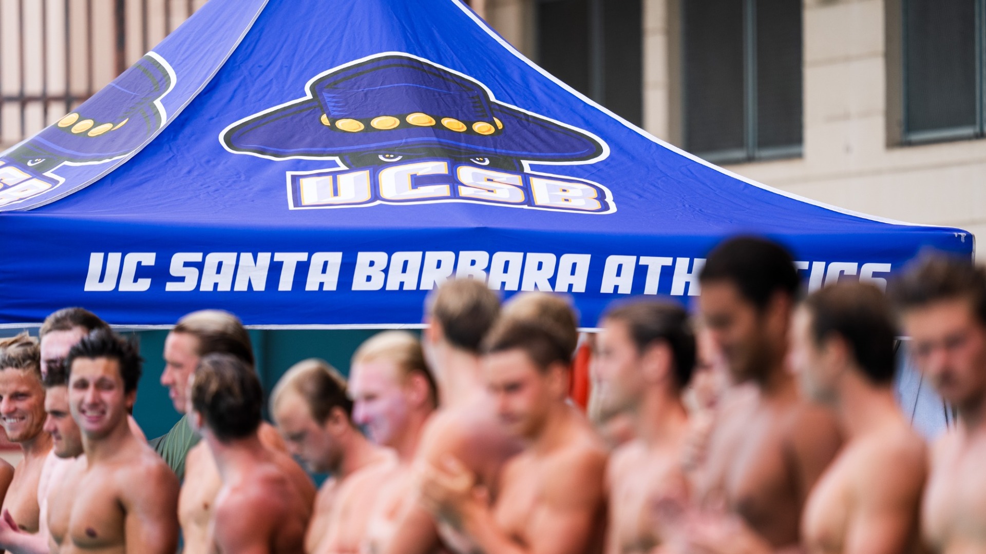 A photo of the Gaucho Men's Water Polo team lined up for introductions. The players are out of focus in the foreground, while the UCSB logo on a blue canopy tent over the team bench is in sharp focus in the background.