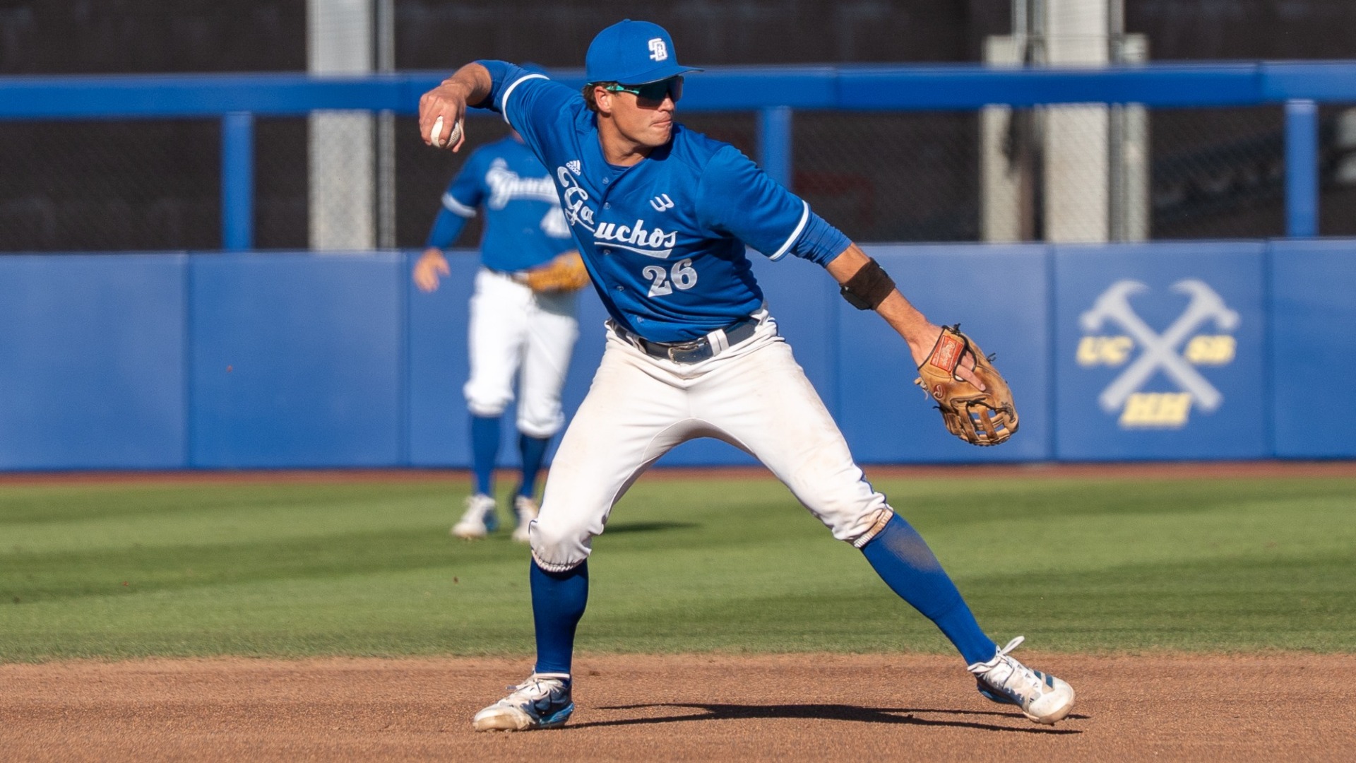William Vasseur (26), wearing the Saturday blue uniform with Gauchos in script across his chest, cocks his right arm back to make a throw after fielding a ground ball at second base. 