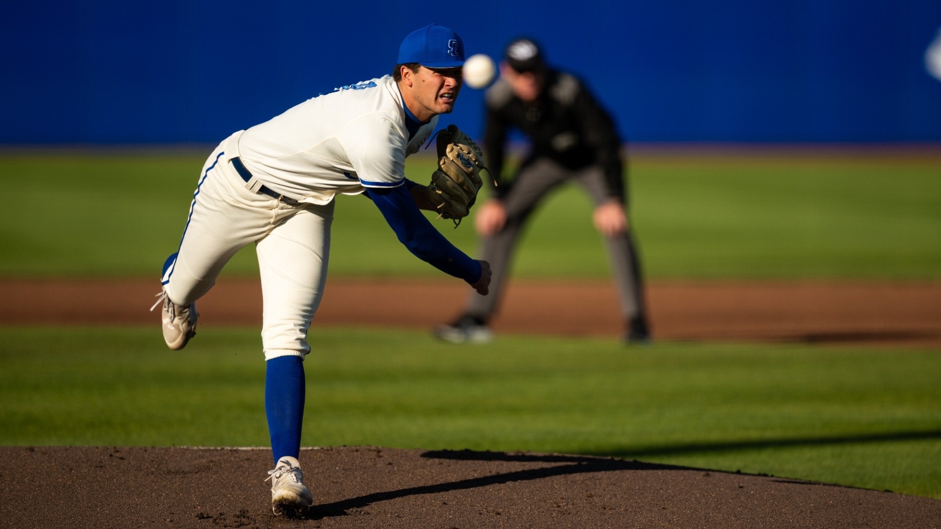 Action shot of Jackson Flora pitching. Jackson, wearing the Gauchos' Friday cream uniform, is landing on his left foot as he finishes his delivery, his right arm now dangling under his torso. The baseball is blurred in the foreground, while a blurred umpire stands in the background.