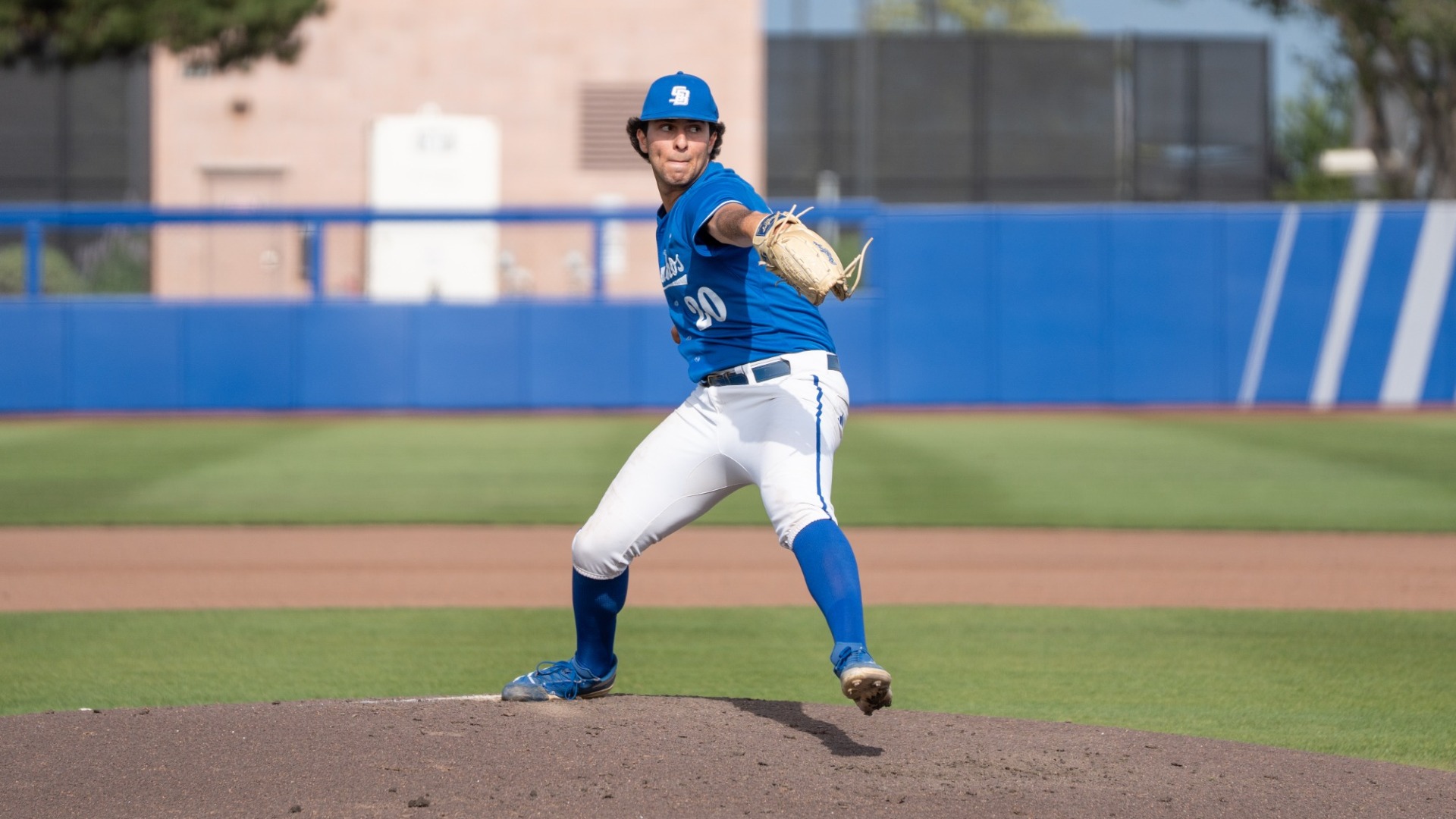 An action shot of Nathan Aceves pitching. Wearing the Gauchos' Saturday blue uniforms, he is stepping into his delivery. His glove hand and left foot are both extended diagonally in front of him. He appears focused as his eyes look toward home plate