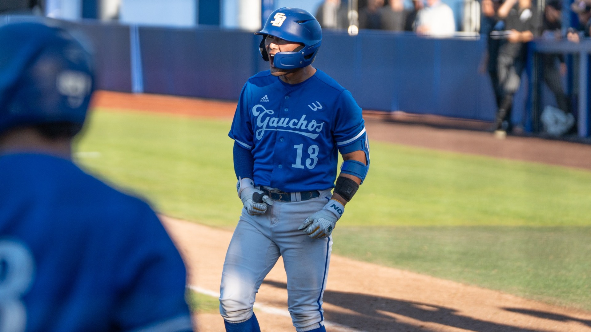 Nico Libed celebrates a Gaucho score. Wearing the Saturday Blue uniforms, we can see him yelling in triumph, even from under the protective face guard on his batting helmet. His hands are down at his hips, with his right hand clenching in a fist.
