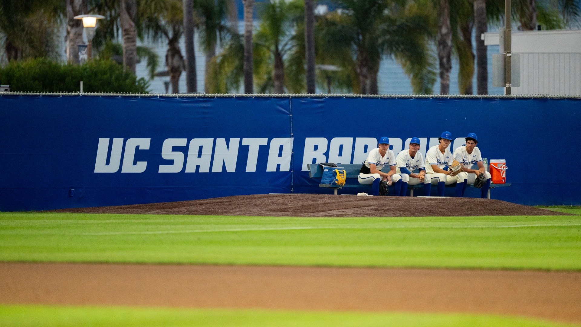AJ Krodel (#45), Chase Hoover (#22), Van Froling (#24) and Stunner Gonzales (#34) sit, hunched, in the Gauchos' bullpen. The four are on a bench next to each other in the right of the frame. Behind them, text on the fence reads UC SANTA BARBARA
