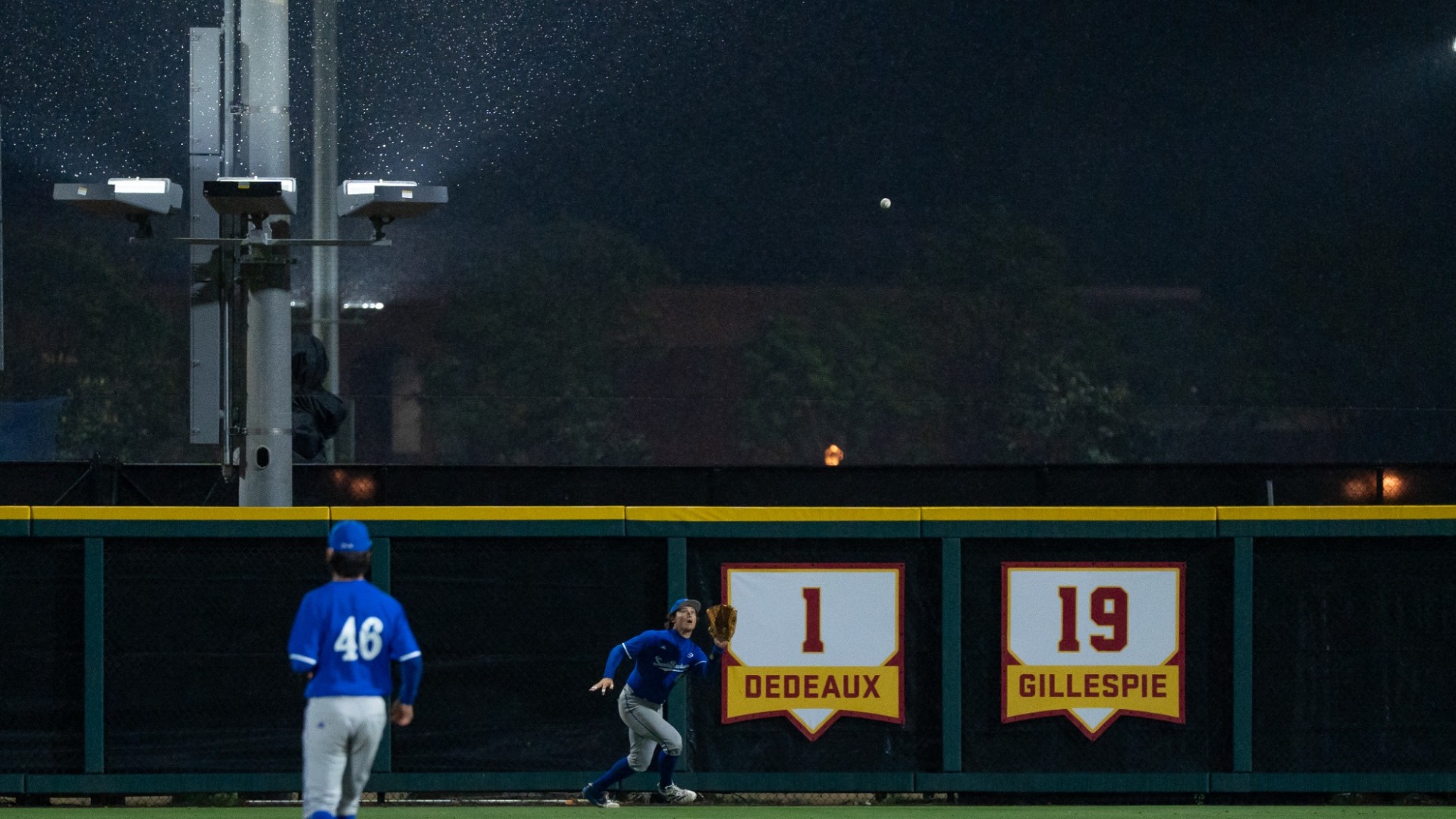 Noah Karliner (#41) prepares to catch a fly ball under a rainy night sky at USC's Dedeaux Field. Behind him are home-plate shaped signs on the outfield wall honoring Rod Dedeaux (#1) and Mike Gillespie (#19)