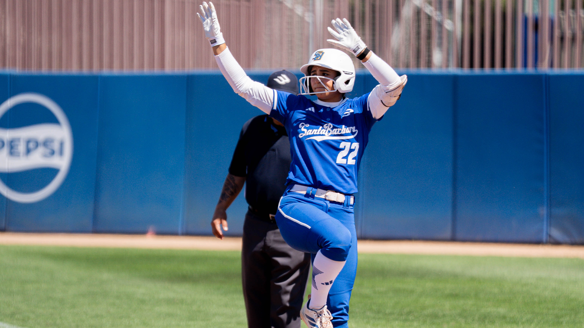 #22 Jazzy Santos celebrates a base hit by making a gesture towards the dugout. She is standing on first base with both hands in the air and one leg in the air at a 90 degree angle.