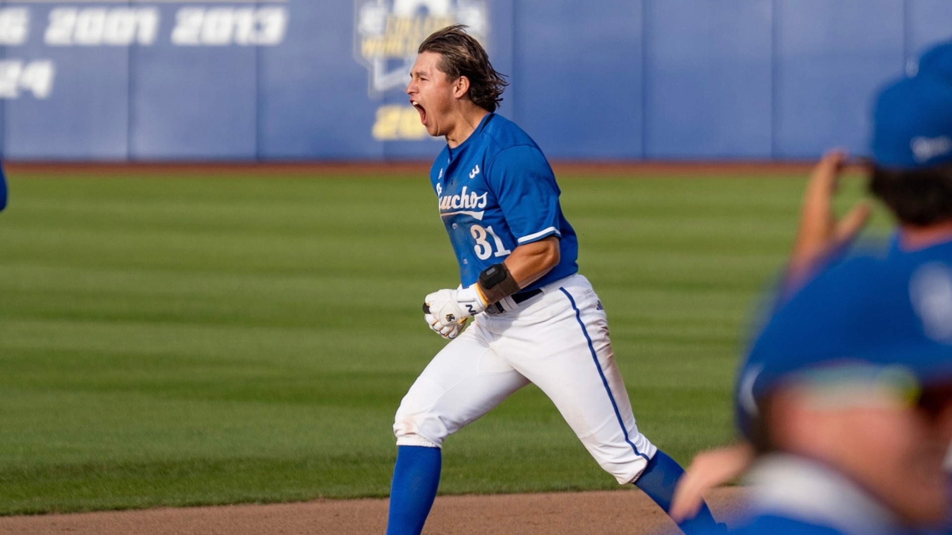 Xavier Esquer (#31) celebrates his walk-off single against UC San Diego. His helmet has flown off and his hair is blown back in the wind as he roars in celebration, pumping his fists in front of him. Out of focus in the foreground are some teammates running to join in his celebration.