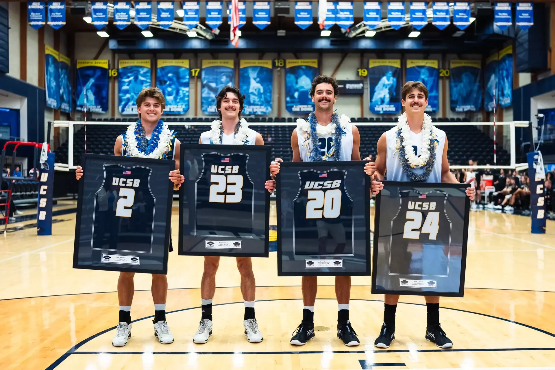 Men's Volleyball seniors Joe Wallace, Ben Pearson, Andreas Schuetz, and Owen Loncar pose in front of the net for a senior night photo. They are holding up framed jerseys and wearing leis. 
