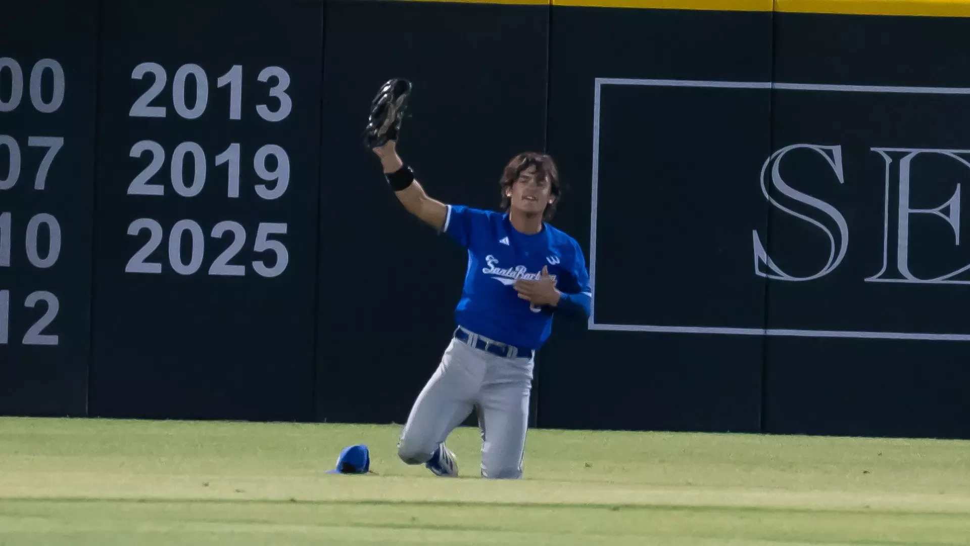 Rowan Kelly (#3) holds up his glove after making a diving catch at UCLA. His cap is strewn on the grass next to him as he sits up on his knees, his right hand raised to show the umpires the ball in his glove. He is beating his chest with his left hand in celebration