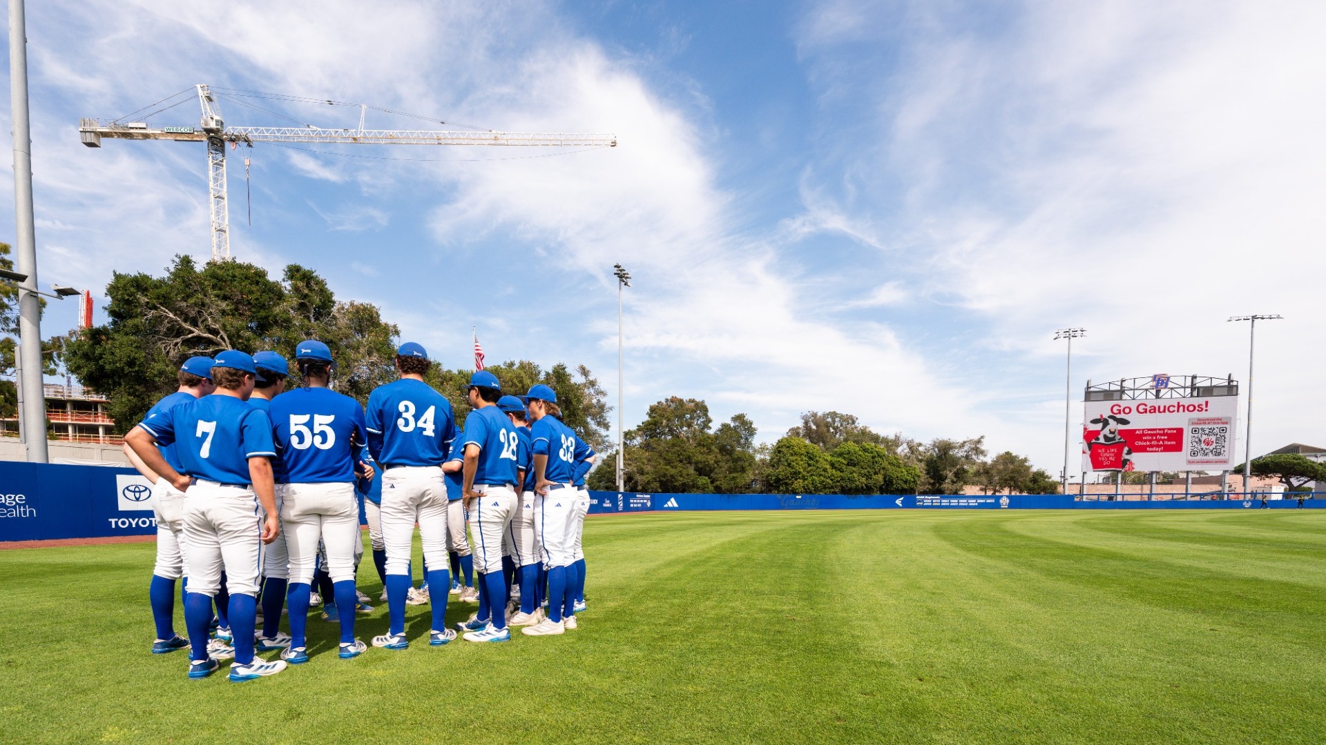 A group of UC Santa Barbara Baseball players huddle together under a blue sky. A large construction crane stands in the background. 
