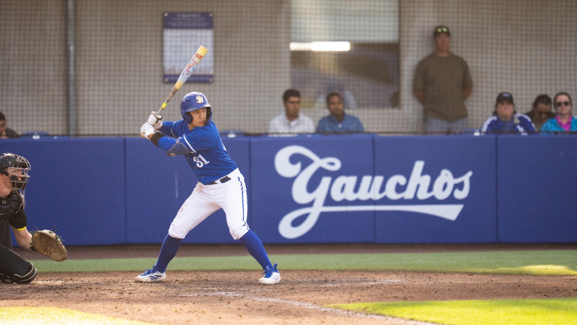 #31 Xavier Esquer, wearing Santa Barbara Baseball's blue uniform, prepares to swing at a pitch in the evening light. The Gauchos' script logo appears on the backstop padding behind him, where some fans watch on.