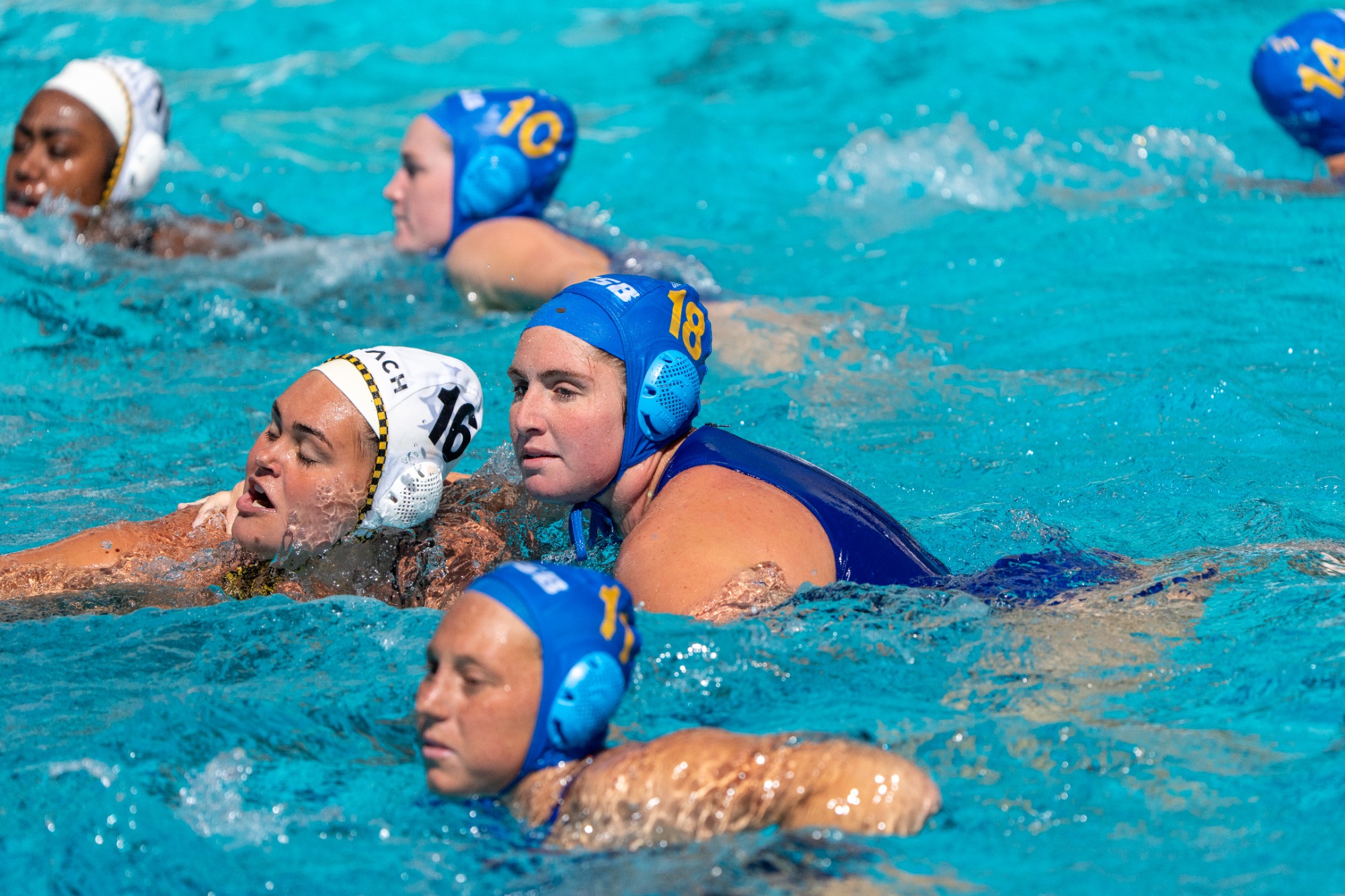 Cami Mras (#18) marking her opponent, Long Beach State's #16, closely from behind. The water around them is busy, with Gaucho teammates out of focus in both the foreground and the background.