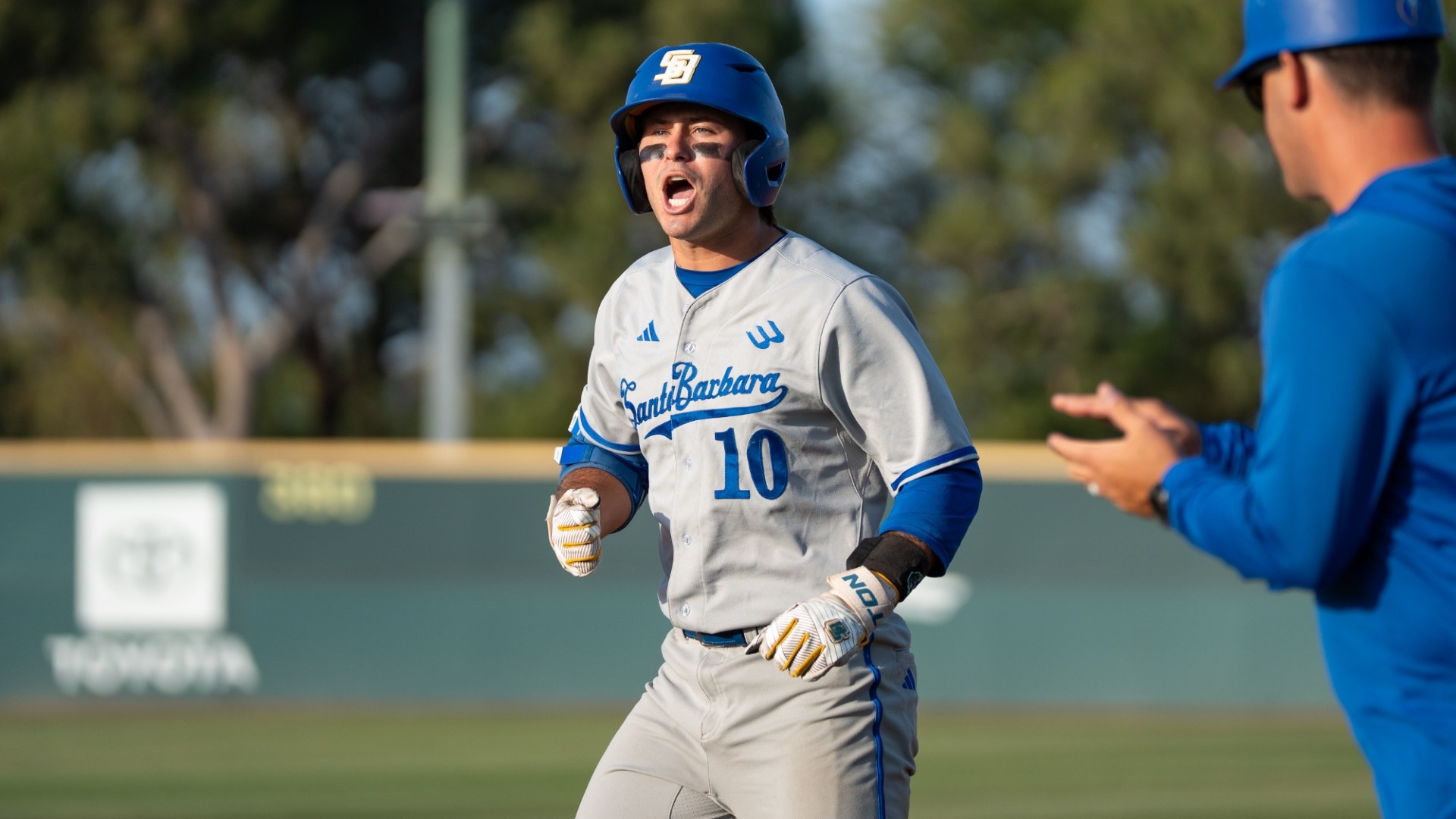 #10 Liam Barrett, wearing the Gauchos' grey away uniform, yells and pumps his fists in celebration. Out of focus next to him, first base coach Kyle Hunt claps his hands